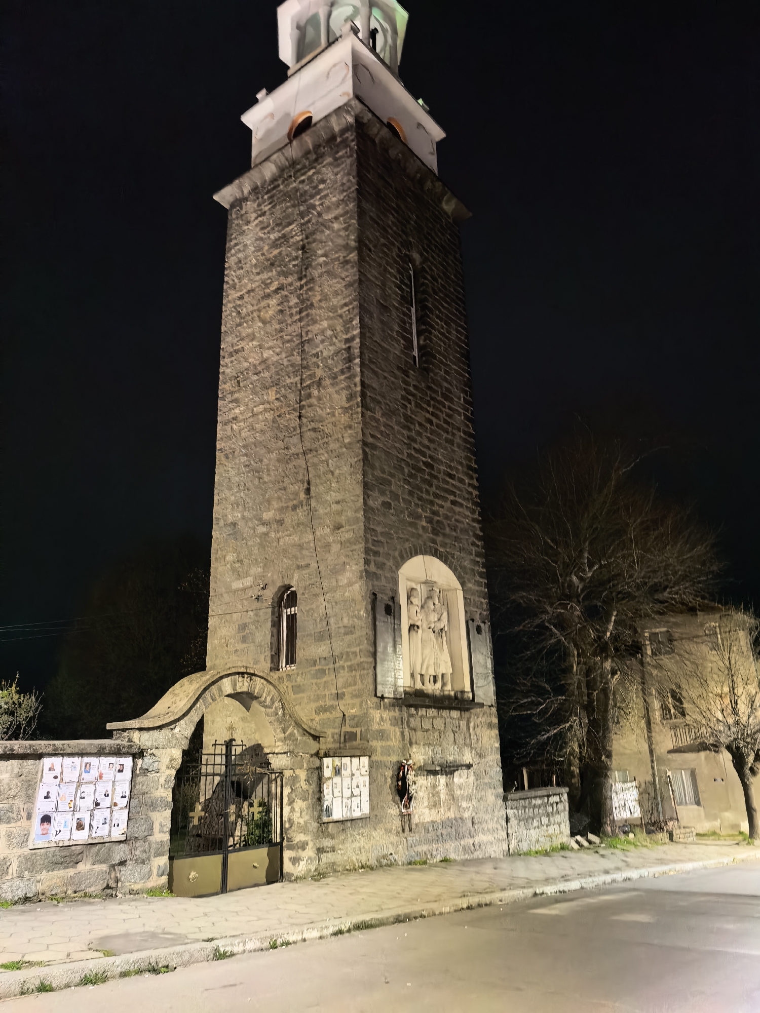 A nighttime shot captures a tall, stone clock tower with an adjacent building in Unknown. The tower, constructed from large stones, rises high into the dark sky and is topped with a white and light-green structure. The tower features an arched entrance gate with black metal doors, and to the left of the entrance, a wall displays numerous posters with faces. Higher up, there is a stone relief depicting three figures. A leafless tree stands next to the tower and the adjacent building. The street is paved, with some vegetation sprouting along the edge. The general atmosphere is subdued due to the dark sky and nighttime lighting.