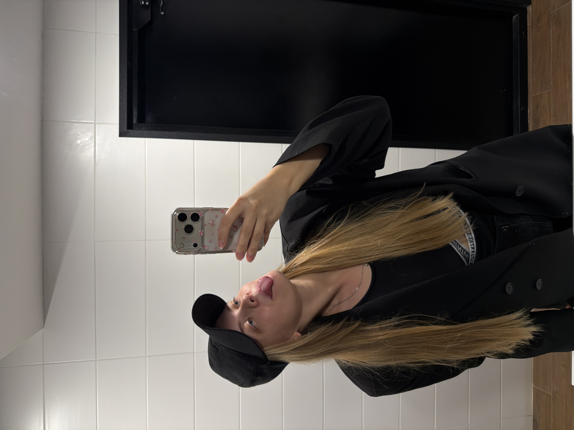 A woman is taking a selfie in a tiled bathroom. The tiles are white. She is wearing a black cap, a black t-shirt, and a black blazer. She is sticking her tongue out. She is holding a smartphone with a clear case decorated with small flowers. Behind her is a black cabinet. The location is unspecified, but based on the prompt, it may be in Izhevsk, Russia.