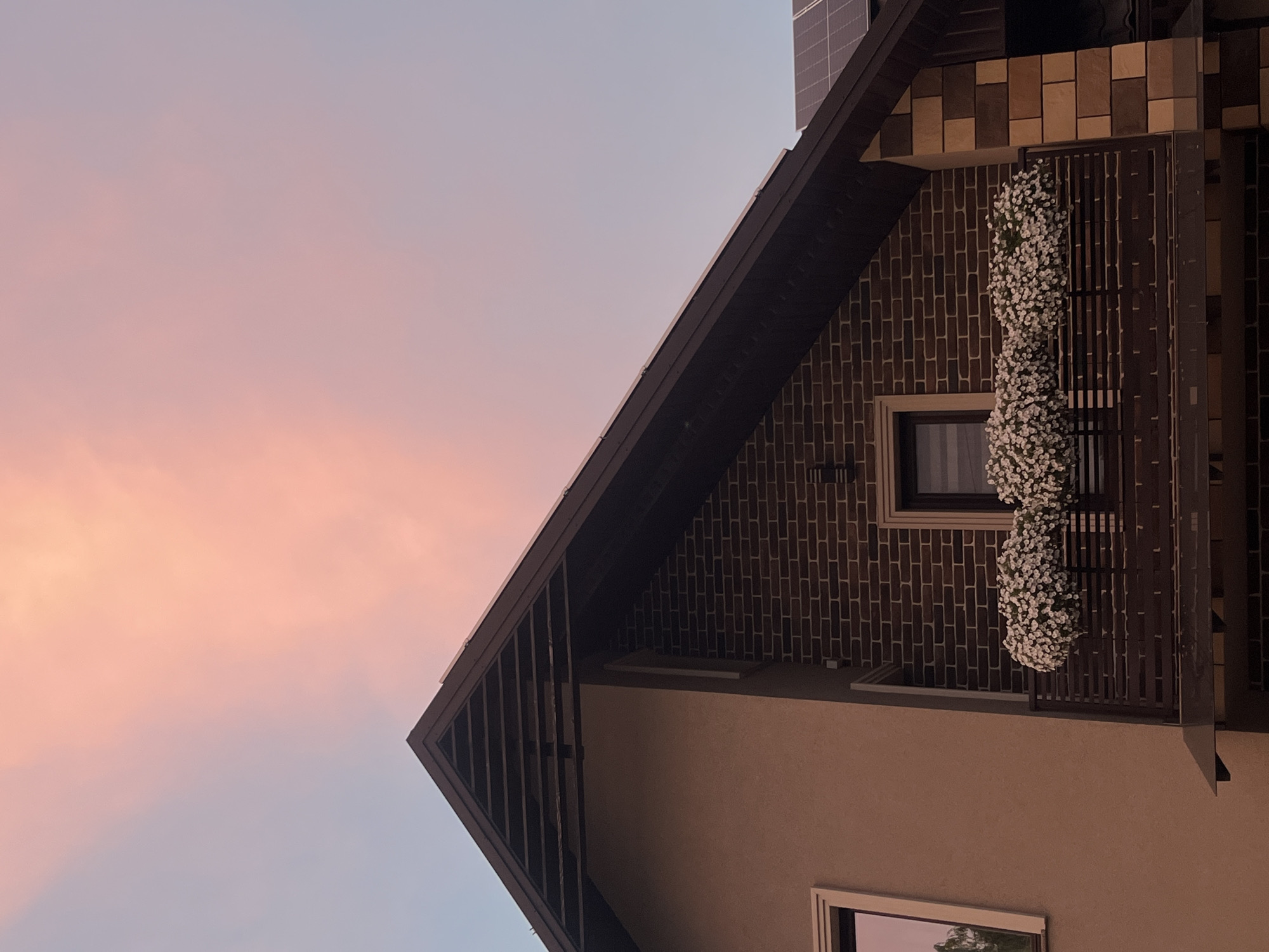 The image shows the upper portion of a house with a brown roof and dark brown brick walls. A balcony with white flowers in planters is visible on the second story. The sky above is a soft pink and blue, suggesting a sunset or sunrise. In the background, solar panels are mounted on the roof. The house appears to be located in a suburban or rural area, with no other buildings or people visible. The photograph is taken from a low angle, looking upwards.