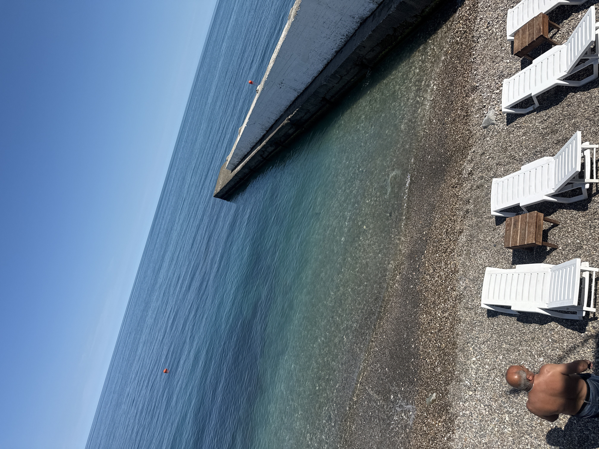 This media file captures a serene, sun-drenched coastal scene on a bright day. The image is taken from an elevated perspective, looking down and across a pebble beach and into the sea.
The clear, vibrant blue sky occupies the upper portion of the frame, meeting the calm, deep blue waters of the sea along a distinct horizon line. The water closer to the shore transitions into clearer, turquoise and green hues, allowing the underlying pebbles to be visible. Ripples are noticeable across the water's surface, indicating a gentle breeze or calm conditions. Two small, bright orange buoys are visible in the water, spaced apart.
A concrete pier or breakwater, light grey on top and darker below, extends from the left side of the frame into the sea, creating a boundary between the open water and the shallower area near the beach.
The right side of the image features a pebble beach, covered in small, dark, and light-colored stones. Arranged neatly on the beach are several white plastic lounge chairs, each accompanied by a small wooden side table.
Towards the bottom right, a person is visible from behind. This appears to be an older man with a bald head and some grey hair on the sides. He is shirtless, wearing dark shorts or swimwear, and stands on the pebbles close to the water's edge, facing away from the camera.
The overall impression is one of relaxation and leisure, typical of a vacation spot. The strong sunlight casts shadows, suggesting it is midday or early afternoon. There is no visible text in the image.