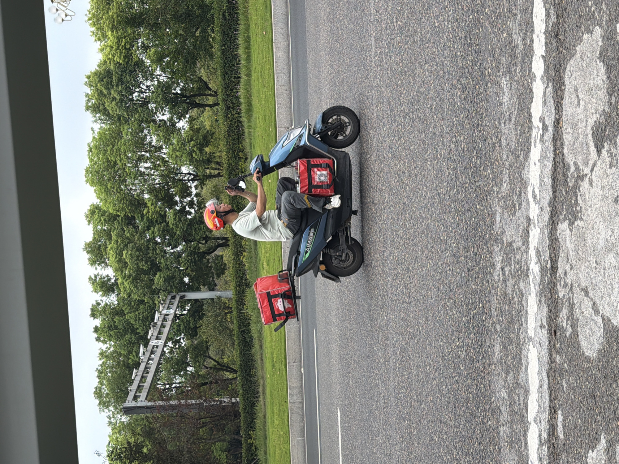 A person wearing a red and orange helmet is riding a blue electric scooter on a paved road. The scooter has two red insulated bags attached, one on the rear rack and one hanging in front of the rider. The rider is wearing a light-colored t-shirt and dark pants, and appears to be holding a mobile phone. In the background, there are trees and a road overhead structure with multiple cameras. The scene suggests a delivery person operating in an urban or suburban environment. The context of Jiangjin District, China, is provided, implying a specific geographical location for this activity.
