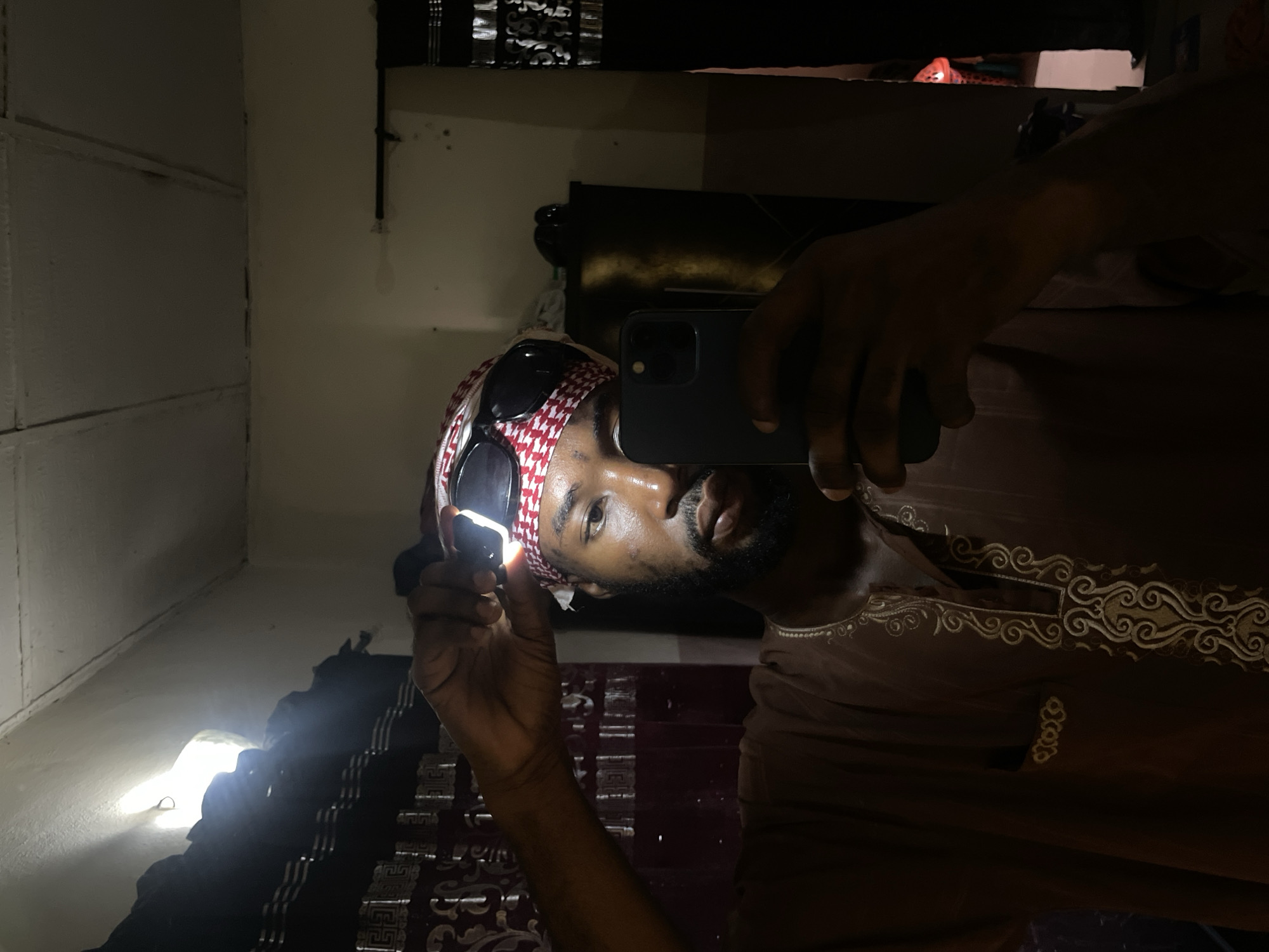 A dark-skinned man, facing the reflection of a mirror, holds a smartphone in his right hand to capture the reflection. He wears a red and white keffiyeh headscarf, sunglasses perched on top of it, and a brown thobe with ornate embroidery around the collar. In his left hand, he holds an unidentified glowing object. The location appears to be indoors, with a wall on the left and some furniture visible in the background. The scene could be in Lokoja, Nigeria.
