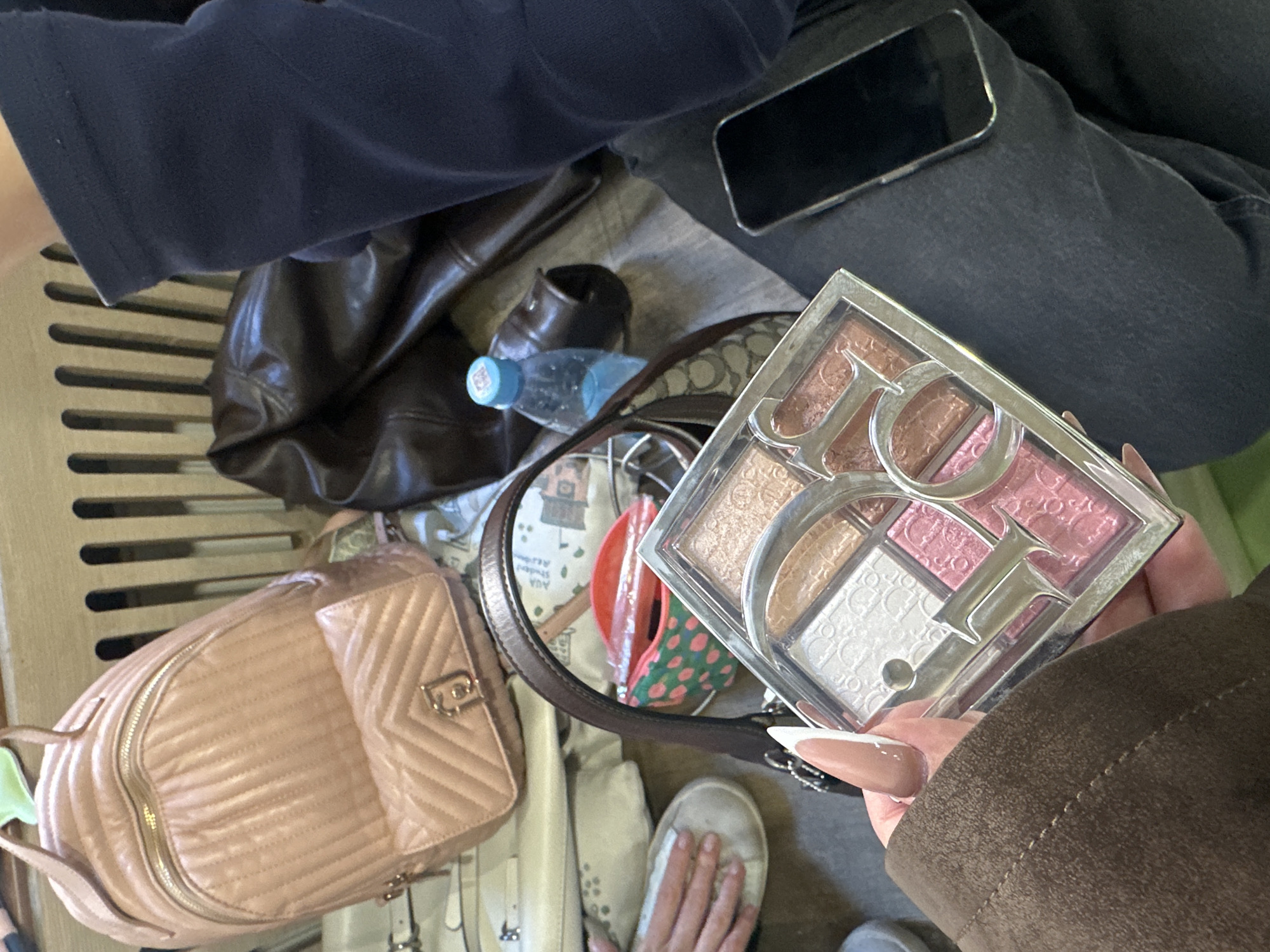 A medium shot shows a collection of personal items on what appears to be the floor of a train or bus, in a casual, somewhat cluttered arrangement. The objects include a pink quilted backpack, a brown leather bag, a clear plastic water bottle, a beige and brown patterned tote bag, a white canvas shoe, and a black smartphone resting on a pair of dark denim jeans. A hand holds a Dior makeup palette. The individual wearing the dark denim jeans is also wearing a dark-colored top. The scene's setting appears to be in Yerevan, Armenia, based on the style of bags and the general environment depicted in the image.