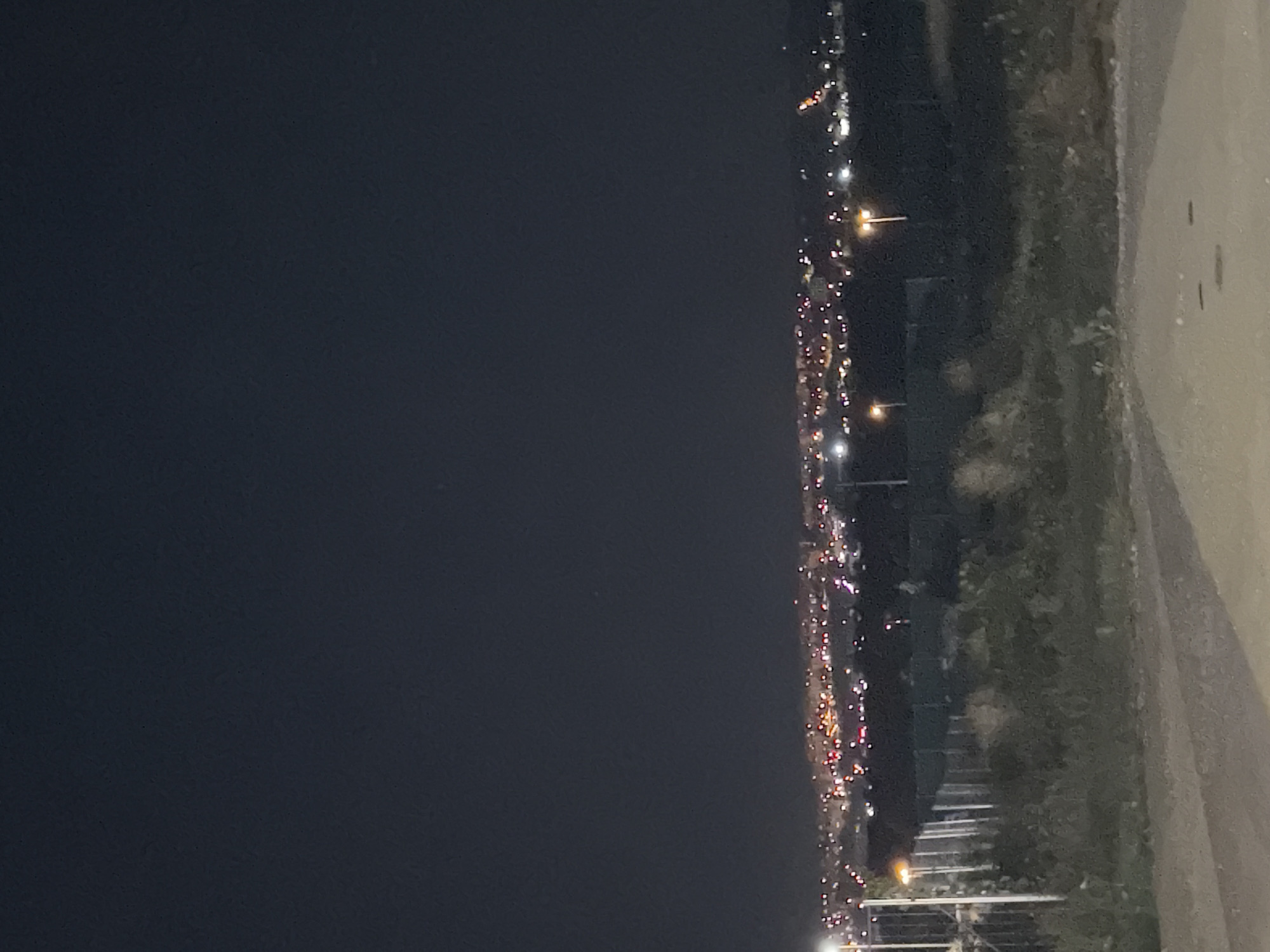 The image depicts a nighttime view of a distant cityscape, likely Nitra, Slovakia, illuminated by numerous lights. In the foreground, there is a dirt path with a few manhole covers. Beyond the path, a fence separates it from a grassy, overgrown area with scattered bushes. Further in the background, streetlights are visible, casting an orange glow on the ground. The cityscape itself is a blur of colorful lights, suggesting a developed urban area. The sky above is dark and clear, with no visible celestial bodies.