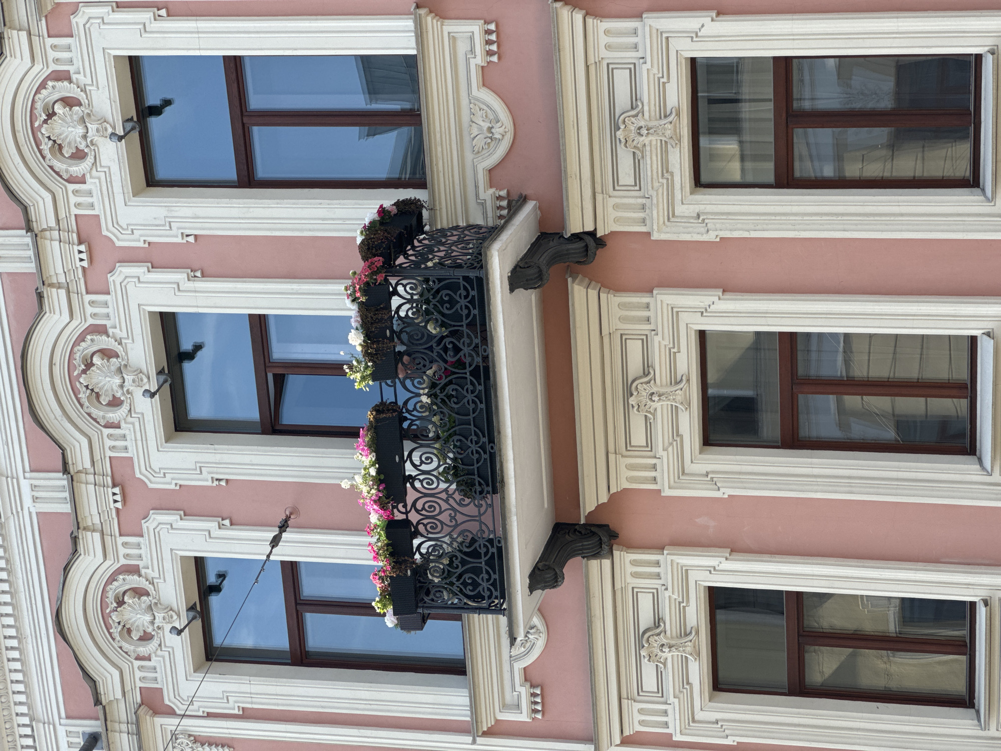 The image shows a close-up, eye-level exterior view of a pink, ornate building facade. Three rectangular windows are visible on the upper level, and three on the lower level. The windows have dark wooden frames and appear to be double-paned, reflecting a blue sky. The upper windows are set within elaborate white architectural moldings that feature decorative carvings and raised trim. Between the upper windows, a small balcony extends outwards. This balcony has a white concrete floor and a black wrought-iron railing with intricate scrollwork. Two black planters filled with pink and white flowers are positioned on the balcony. Below the balcony, a large white ornate molding runs horizontally across the facade, with decorative elements mirroring those around the upper windows. The lower windows are also framed by similar white architectural moldings, though they are less elaborate than those on the upper level. The building's pink stucco exterior is visible between the architectural details.