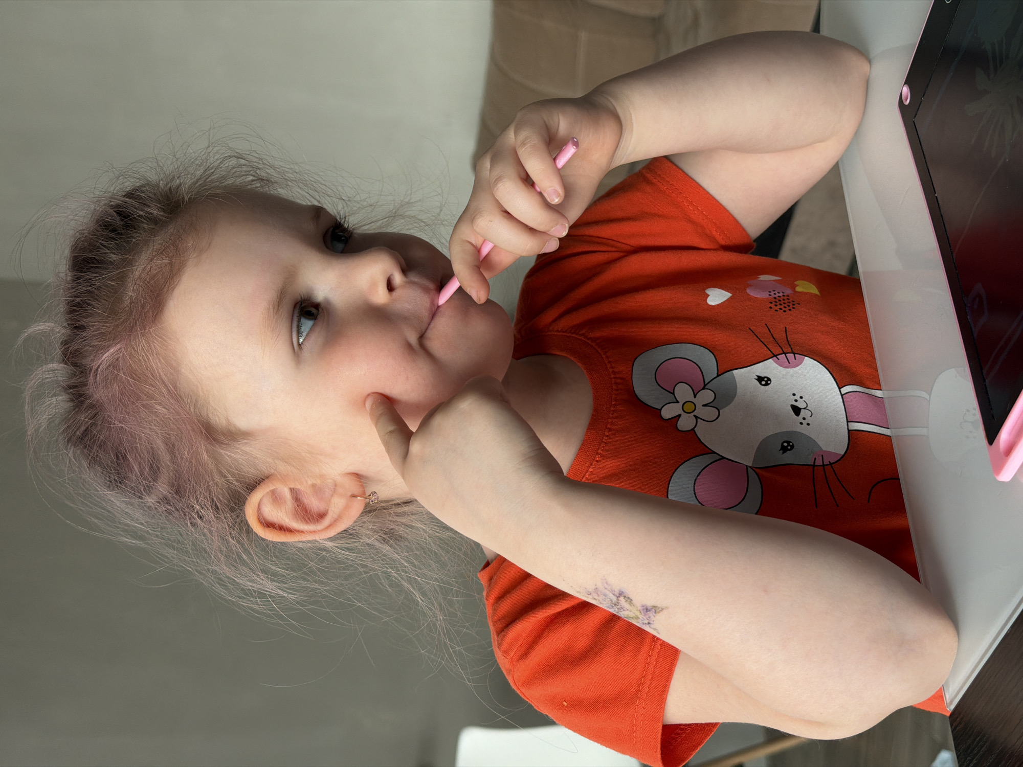 A young child with fair skin and light, slightly tinted pink hair is seated at a white table. She is wearing an orange t-shirt with a mouse and rabbit graphic and an earring in her left ear. She has a purple floral temporary tattoo on her left forearm. The child is touching her cheek with her left forefinger and holding a small pink stylus in her right hand, partly in her mouth. Beside her on the table is a dark-colored electronic drawing tablet with a pink frame. The background includes a gray wall and a beige cushioned object. The location is in the city of Chaikovsky, Russia.