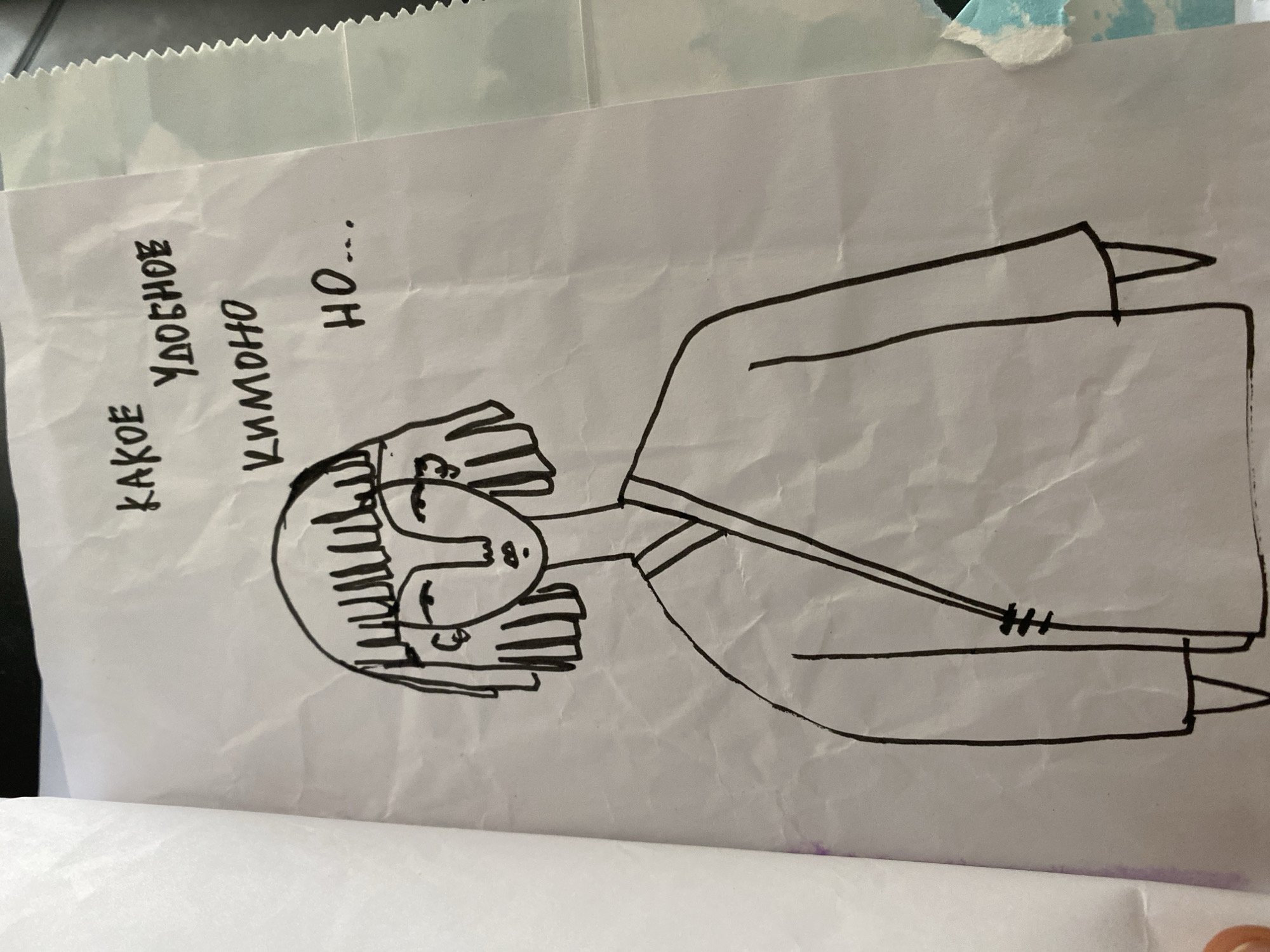 A black ink drawing on white paper depicts a figure wearing a kimono. The figure is centered on the page, facing left. The figure has a round head with stylized vertical lines representing hair. The face is simply drawn with a straight nose and rectangular eyes. The kimono is loosely sketched with long sleeves and a rectangular body. Russian text, “КАКОЕ УДОБНОЕ КИМОНО, НО…” is written vertically to the left of the figure. The paper appears to be slightly crumpled. There is no discernible indication of the location of Lâm Viên - Đà Lạt, Vietnam, nor any indicators of a city location in the scene.