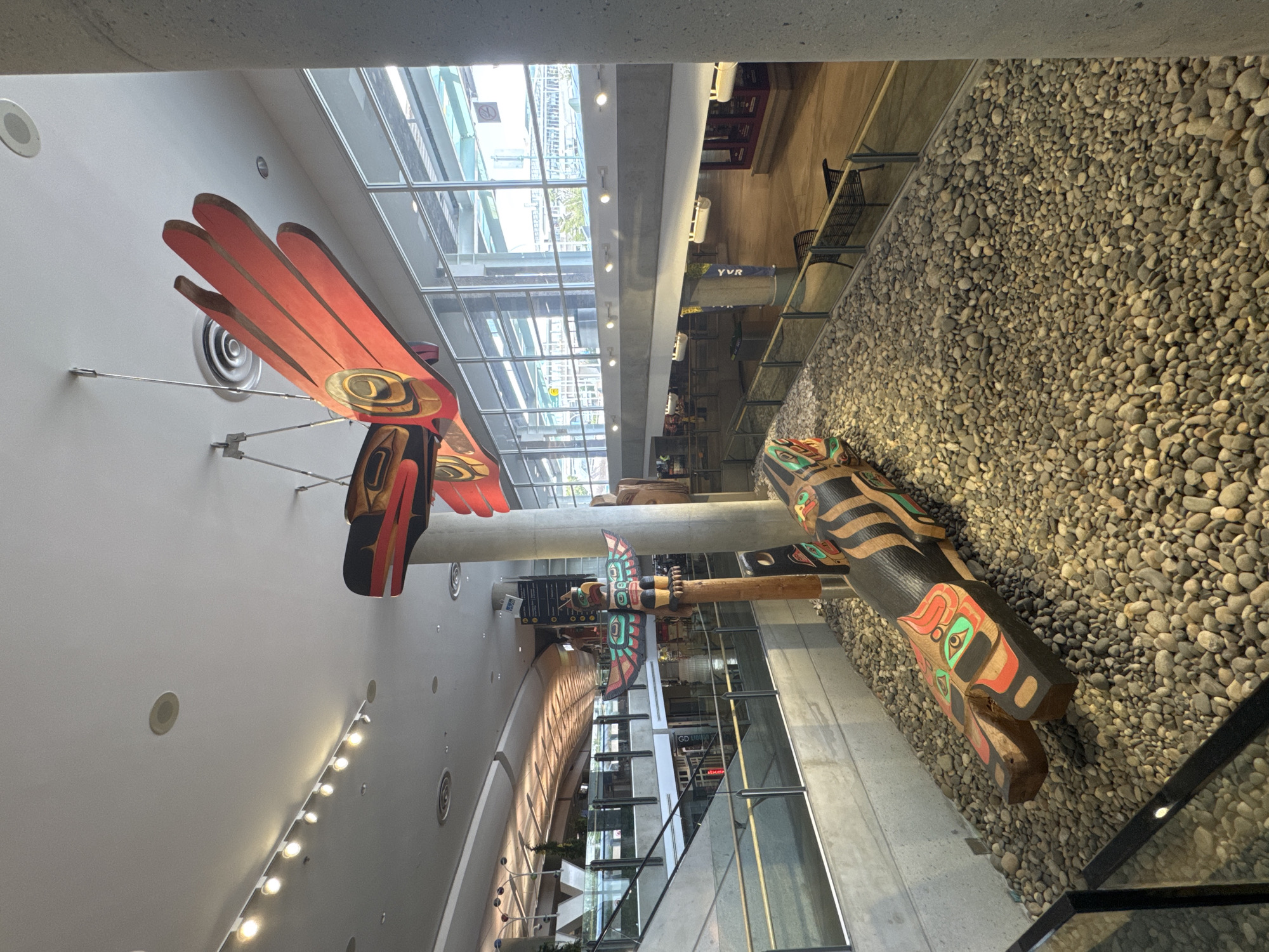 The image depicts the interior of Vancouver International Airport (YVR) in Richmond, British Columbia, Canada. Visible are several large, ornately carved Indigenous totem poles. The totem poles are mounted and displayed within the airport terminal. One totem is topped with a wooden carving of a bird. The background includes glass walls and open walkways. The right side features a wall covered in small gray stones.