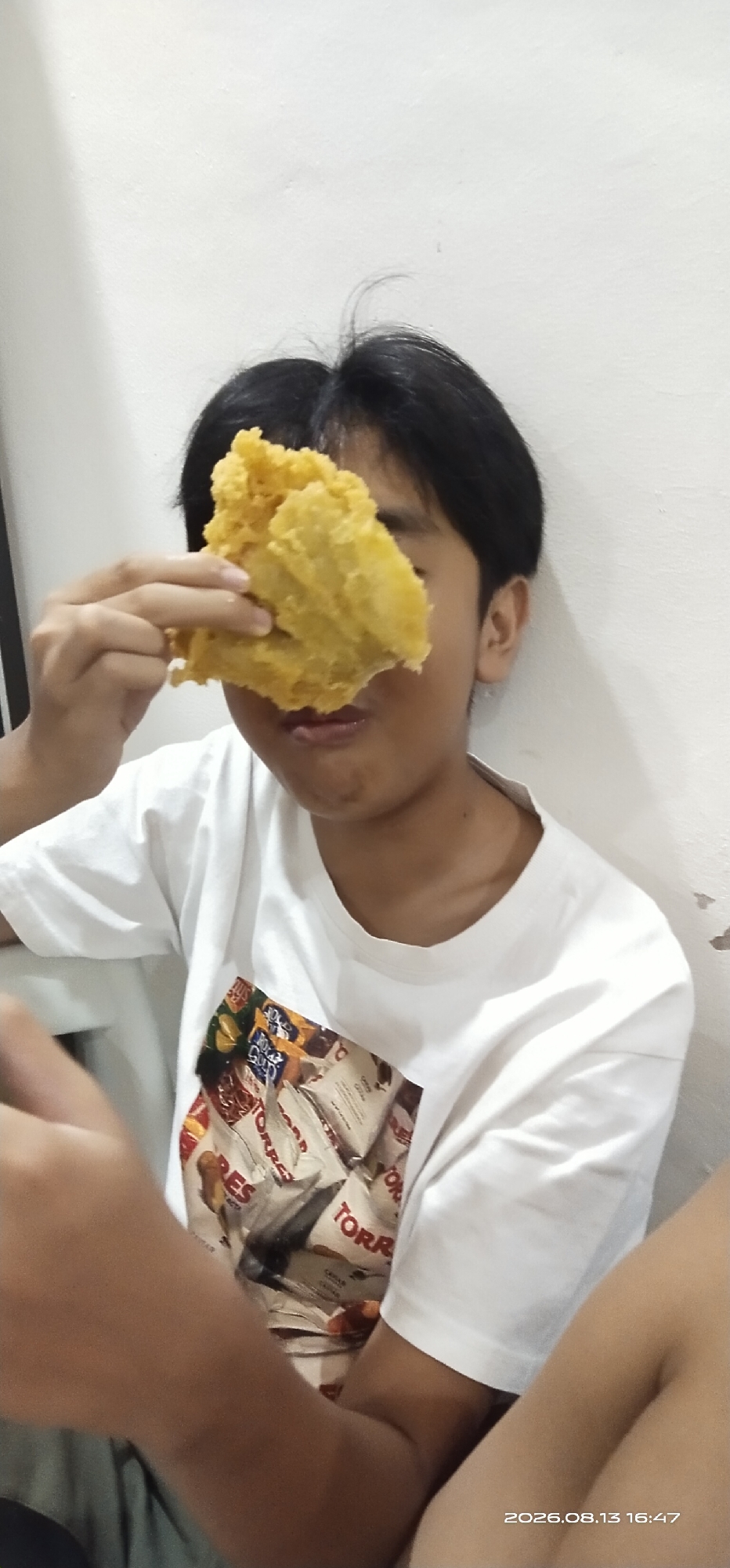 A young person with dark hair wears a white t-shirt featuring a collage of food packaging. They hold a piece of fried food in front of their face, obscuring one eye and part of their mouth. The background is a plain, off-white wall. The scene appears to be indoors, possibly in Matabu, Indonesia, given the date stamp "2026.08.13 16:47" in the lower right corner.