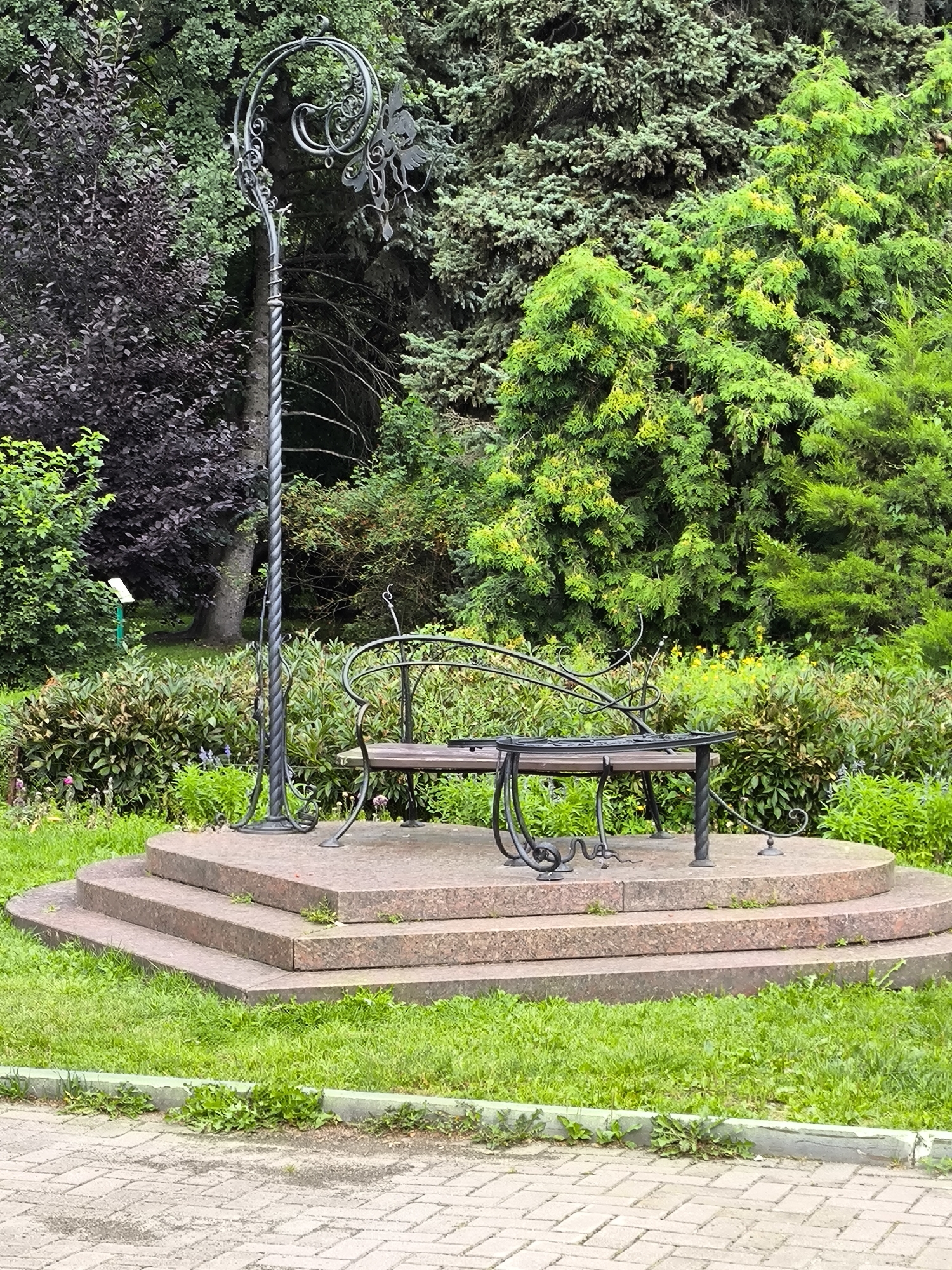 A decorative park bench with a matching ornate lamp post is situated on a tiered, circular granite platform in a park. The bench is made of dark metal with wooden slats. The lamp post features intricate metalwork at its top. The platform is surrounded by lush green grass, a paved walkway, and various trees and bushes. In the background, a dark-leaved bush is visible to the left, and dense evergreen trees dominate the right side. A small white sign on a green pole is also present in the background to the left. The overall scene is a well-maintained outdoor public space, likely in Yekaterinburg, Russia, given the context. There are no people or animals visible in the image.