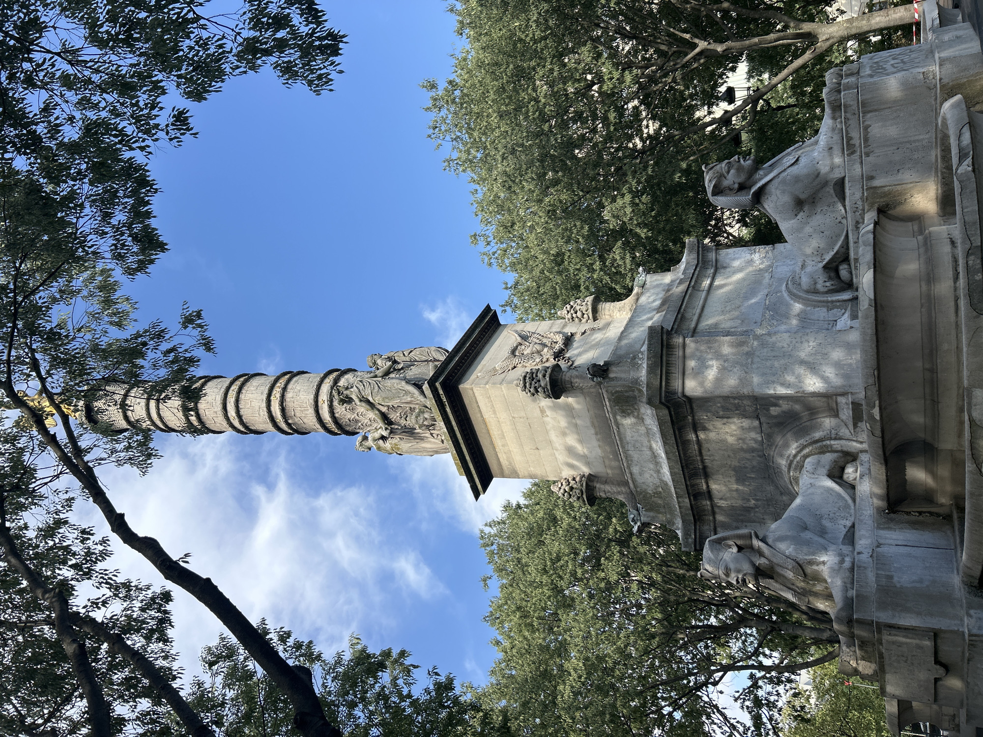 The image shows the Fontaine de lausnya in Paris, France. The monument is a stone column topped with figures and sphinxes at its base. The monument is surrounded by lush green trees and a bright blue sky with scattered clouds. The sun is shining, casting shadows on the monument and trees. The monument appears to be made of weathered stone, suggesting it is an old historical structure. The scene is tranquil and serene, showcasing a beautiful Parisian landmark.
