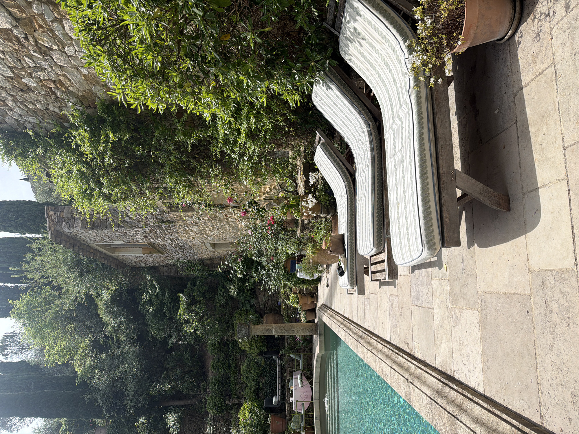 This media file presents a sun-drenched outdoor scene depicting a luxurious poolside area in Saint-Victor-la-Coste, France. The primary focus is a clear, turquoise swimming pool with a light-colored stone-paved patio surrounding it. Along the pool's edge, three wooden sun loungers with striped white and light grey/green cushions are neatly arranged, casting distinct, long shadows that indicate it is likely mid-morning or early afternoon under bright, clear skies.
The setting is characterized by abundant natural beauty and traditional architecture. A large stone building, possibly a historic residence, stands in the background on the left, its walls partially obscured by dense, vibrant green foliage and climbing plants. Various trees, including tall, slender cypress trees on the far left, contribute to the lush, verdant environment. Stone walls, heavily integrated with greenery, define the property's boundaries.
Further into the scene, a smaller poolside seating area is visible, featuring a table draped with a pink tablecloth and two chairs, suggesting a tranquil spot for dining or relaxation. A decorative stone column stands nearby. Potted plants, including one terracotta pot with white flowering blooms beside the loungers, add touches of floral charm. The overall impression is one of serene luxury and a peaceful retreat, showcasing the beautiful natural and architectural elements typical of the French countryside. No people or visible text are present in the image.