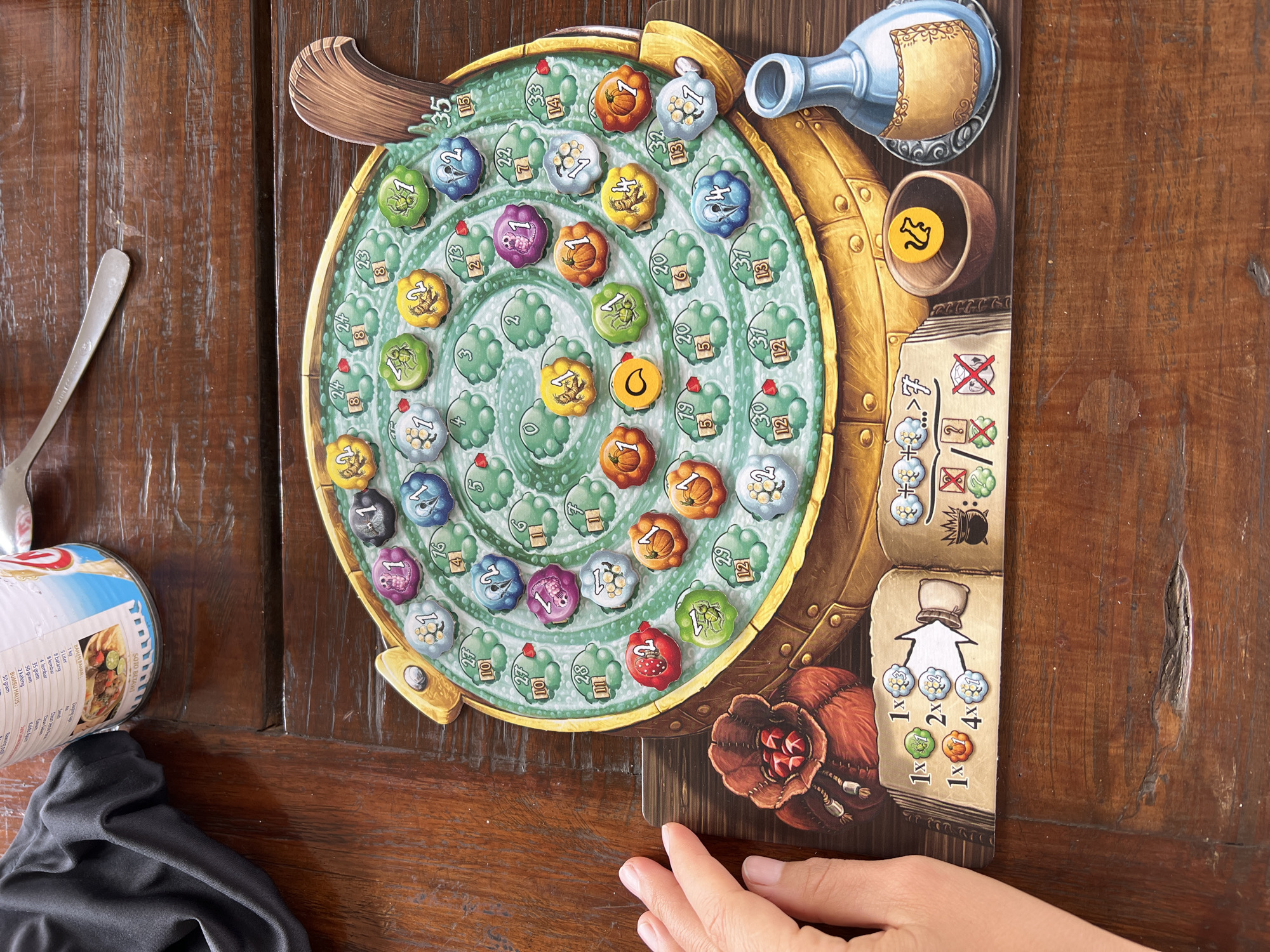 This is a close-up, eye-level shot of a board game being played on a wooden table. The game board is circular with a spiral pattern and is filled with small, round tokens of various colors and designs, some featuring numbers and symbols. One token shows a yellow chicken on a black background. The board is placed on a dark brown wooden table with visible grain and joins.
In the foreground, on the left, a person's hand with manicured fingernails is visible, resting on the edge of a cardboard game accessory. This accessory is a flat, rectangular piece with illustrations of game tokens and symbols, indicating game rules or player information. To the right of the board game, a small, wooden bowl is visible containing a single yellow token with a black chicken symbol. Next to the bowl is a blue glass bottle with a gold label, also part of the game. In the upper left corner, a black fabric item, possibly clothing, is partially visible, along with a can of "Soto Betawi" soup.
The image does not provide any information about the location in the city of Busungbiu, Indonesia. The scene is focused solely on the board game and its immediate surroundings on the table.