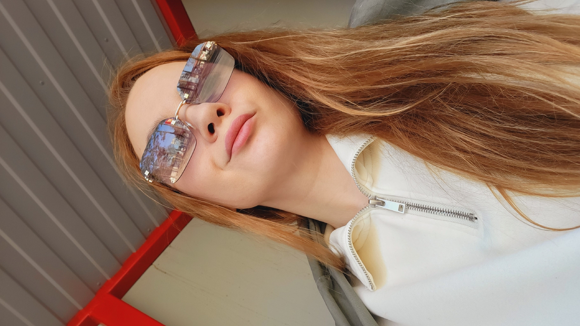 A medium shot shows a woman with long, reddish-blonde hair wearing sunglasses and a white zip-up jacket. The reflection in her sunglasses shows buildings and trees, suggesting an urban environment. The background includes a gray and red horizontal striped structure, likely a building or part of an outdoor setting. The photo seems to be taken at an angle, looking up towards the subject. Without additional information, pinpointing the exact location in Chaikovsky, Russia is impossible.