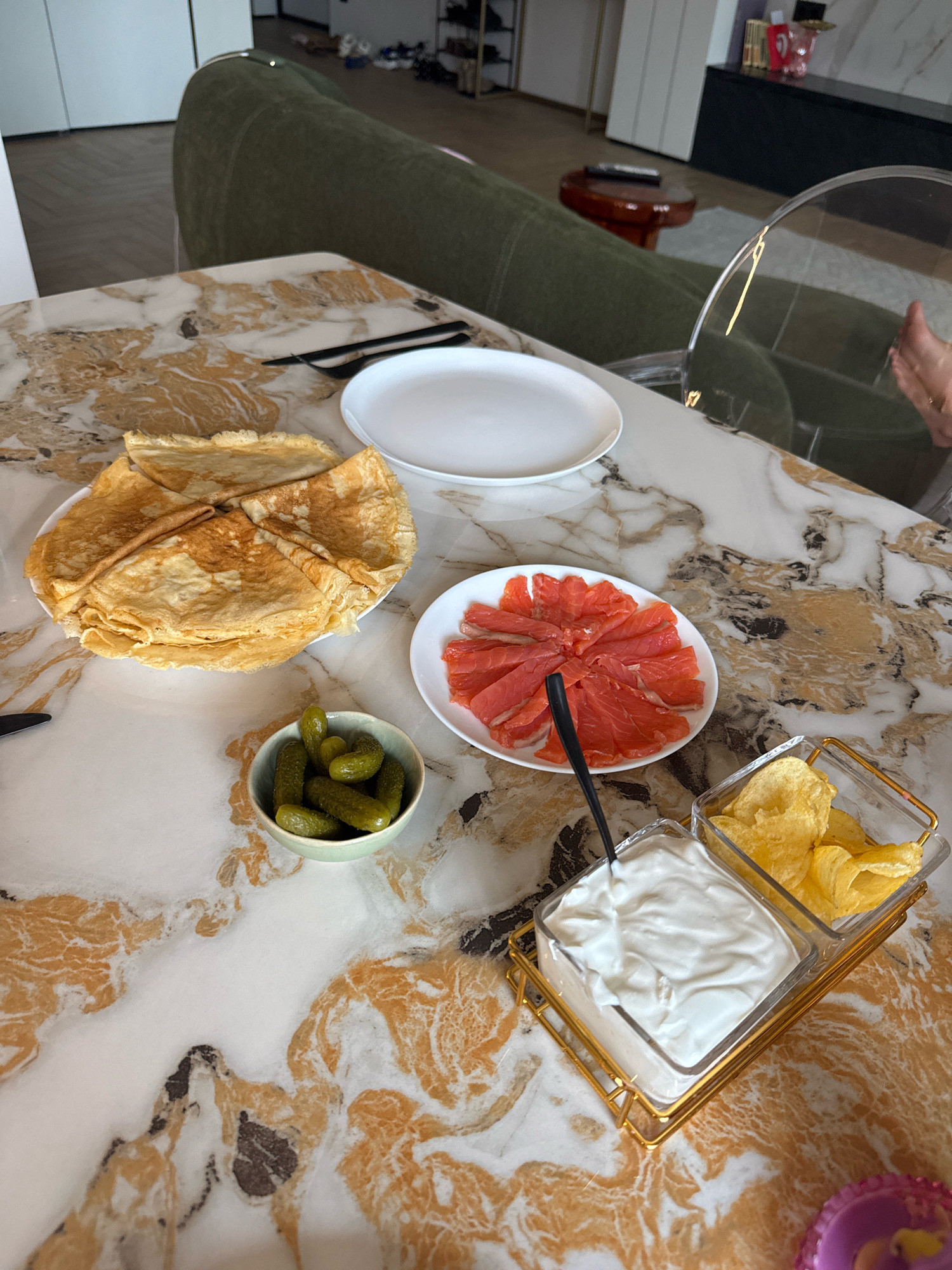 The image shows a table set for a meal, likely in a modern apartment in Rostov-on-Don, Russia. The table has a marble-patterned surface. On the table, there is a plate of crepes, a plate of sliced smoked salmon arranged in a circular pattern, a small bowl of gherkins, and a clear dish divided into two sections, one filled with a white creamy dip (likely sour cream or yogurt) and the other with potato chips. There is also a white dinner plate to the left of the crepes, and a black fork. In the background, a green couch is visible, along with furniture and what appears to be a closet. A person's hand is partially visible on the right side of the frame, near a clear chair. The lighting suggests an indoor setting.