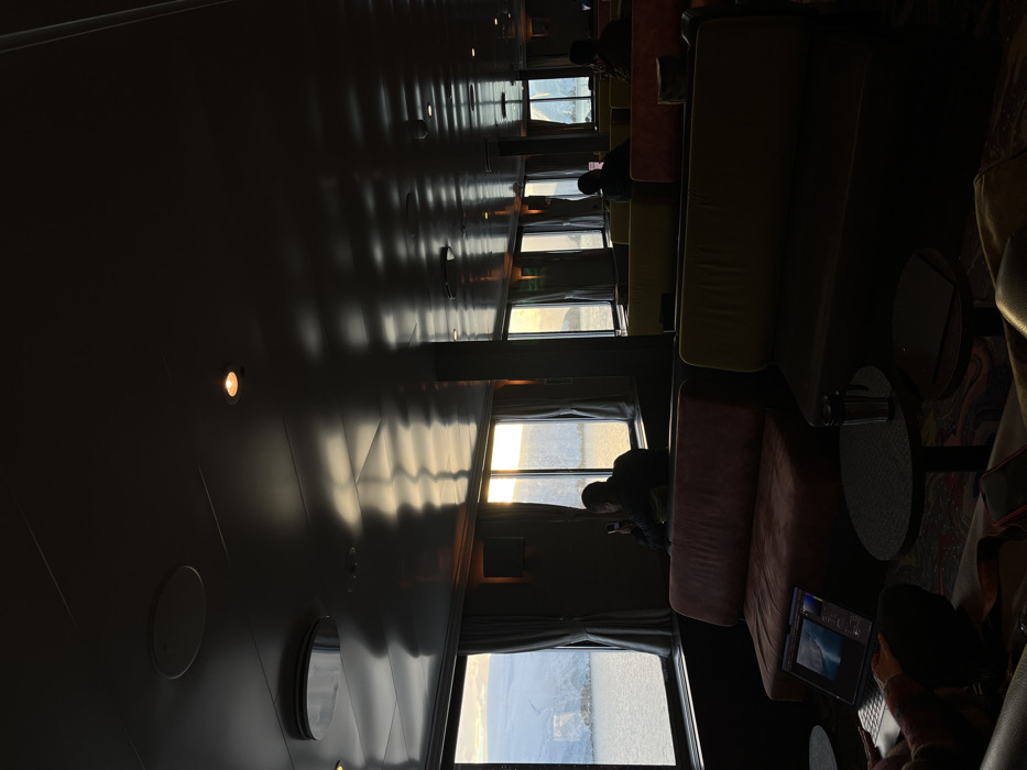 The image shows an interior with a row of large windows offering a view of a bright, hazy landscape. Along the windows, there are sofas where people are sitting. One person is working on a laptop. The ceiling is dark and glossy, reflecting the light from the windows. The room is dimly lit, with the light mainly coming from the windows.