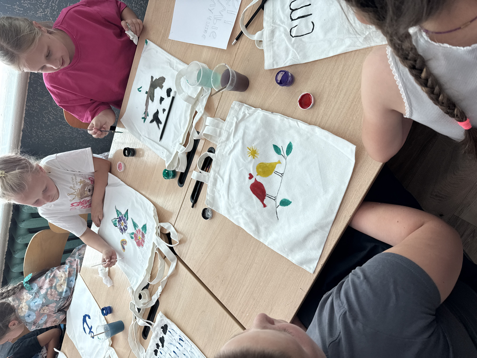 An overhead medium shot captures a group of young children and an adult participating in a painting activity at a wooden table, likely indoors in a classroom or workshop setting in an unknown location. The focus is on the white canvas tote bags laid out on the table, adorned with various painted designs. In the foreground, a child's arm, visible from the shoulder down, rests on the table, with a partial view of a white tote bag featuring a painted design of two birds on a branch. Adjacent to this bag are small containers of red and purple paint. To the right, another white tote bag has a design of two large black letters "cm" painted on it, and a small container of blue paint is visible nearby. Further up the table, a girl in a pink t-shirt is actively painting a design on a white tote bag with a paintbrush, holding a white cloth in her other hand. Next to her is another girl in a white t-shirt, also painting on a white tote bag with a floral design, holding a paintbrush and a cup of blue liquid. Other tote bags with painted designs, including an anchor and a rain pattern, are visible on the table, along with various paint supplies such as brushes, paint containers, and cups of colored liquid. The children are engaged in the creative process, with the adult likely supervising. The overall scene suggests a fun and educational art session.