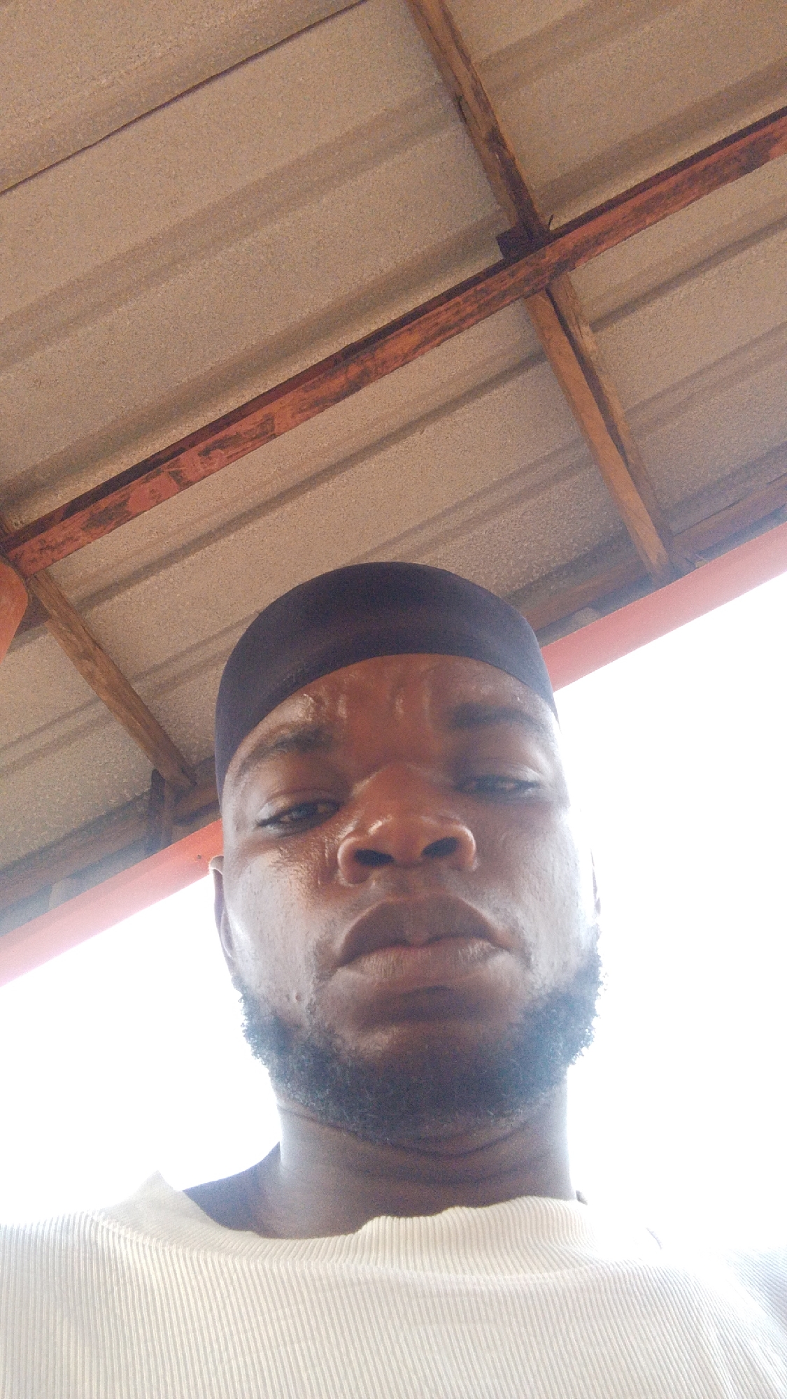 The image is a low-angle selfie of a man with dark skin, a closely cropped beard, and a black head covering. He appears to be perspiring. He is wearing a white, ribbed shirt. Above him is a roof constructed of corrugated metal panels supported by rusty metal beams. The photograph was taken in an outdoor setting with bright, diffused light. The location is speculated to be the city of Enugwu-Ukwu, Nigeria.