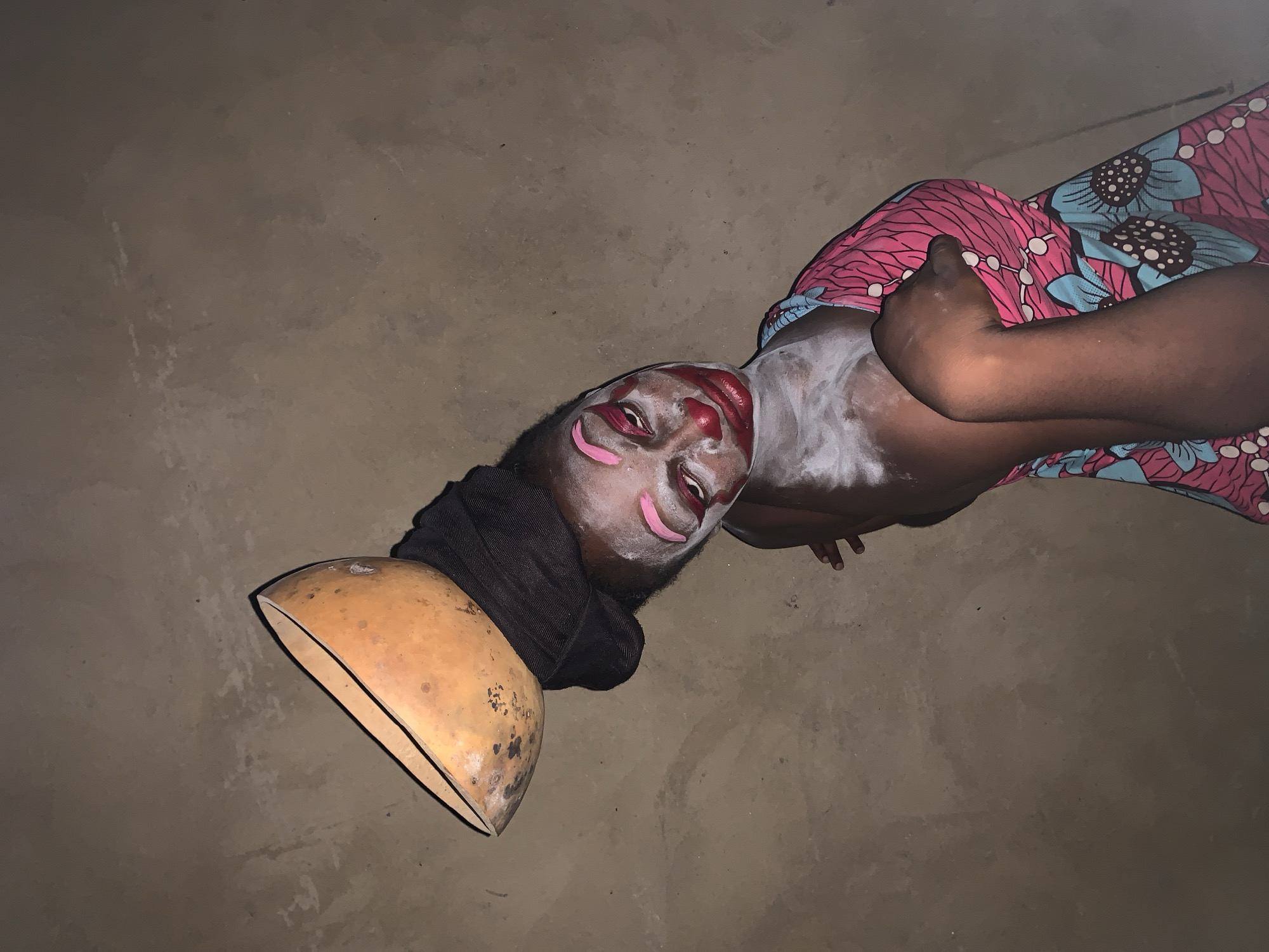 In Agban, Nigeria, a person adorned with clown-like makeup and a gourd on their head is depicted. The individual has white powder on their face and chest, with pink and red makeup applied to their eyes, nose, and lips, creating a theatrical appearance. They are wearing a vibrant pink and blue patterned dress and a black head wrap. A light brown, hollowed-out gourd rests on top of the head wrap. The background appears to be a textured, earthen wall, suggesting an outdoor or rustic indoor setting. The lighting is somewhat dim, casting shadows and highlighting the makeup and the gourd. The person's pose is dynamic, with their head tilted and an arm resting on their chest.