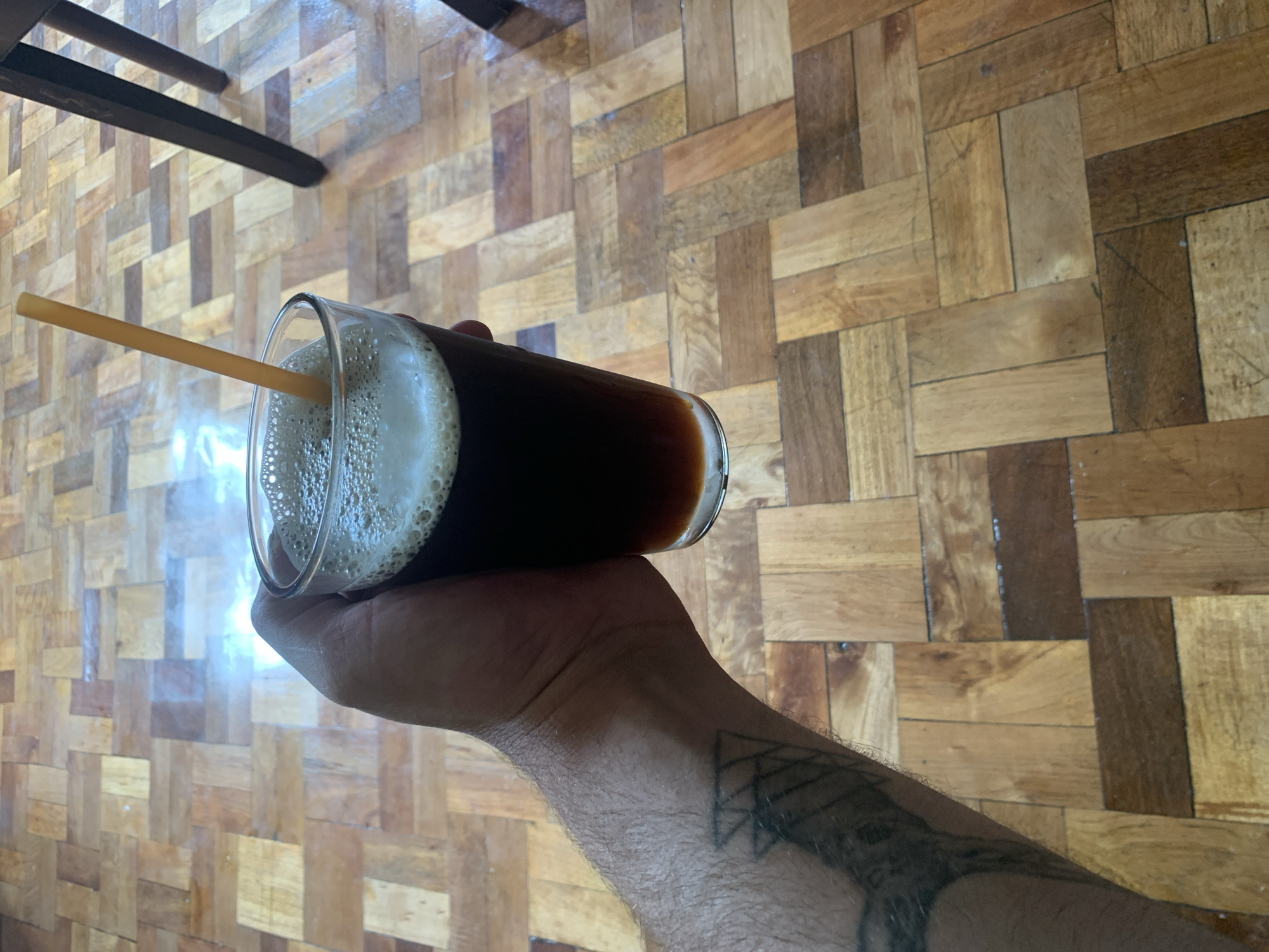 A hand is holding a glass of dark liquid with a straw, resting on a parquet floor. The hand has a tattoo on the forearm and wrist. The glass contains a dark beverage with foam on top, and a straw is inserted into it. The floor is made of wood planks arranged in a parquet pattern, with varying shades of brown and tan. In the background, the legs of a dark wooden chair are visible. The scene is indoors. There is no information in the image to suggest the location is in Chiatura, Georgia.