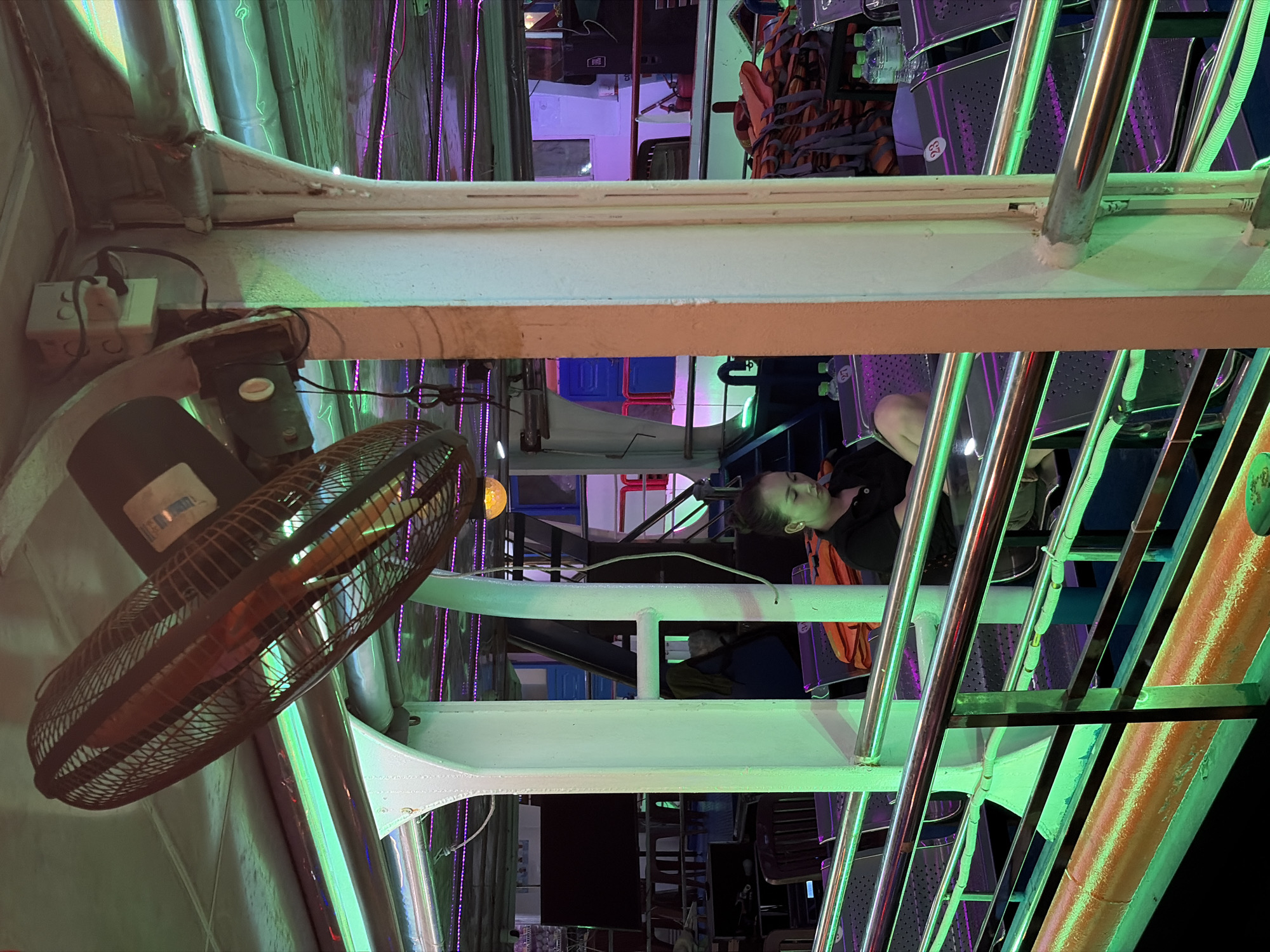 This image captures the interior of what appears to be a boat or ferry at night, illuminated by vibrant artificial lighting. The scene is likely a leisure cruise, given the colorful ambiance and the setting of Đà Nẵng, Vietnam.
In the foreground, a large, dark-colored electric fan with a protective metal cage is suspended from the ceiling. Adjacent to its mounting, a white power outlet with several plugs is visible, connected to wires. The boat's structure consists of sturdy white and light-colored metal beams and railings, which are highly reflective of the neon-green and purple LED strip lights that run along the ceiling and walls.
At least one young person with dark hair, wearing a dark top and an orange life vest, is visible, looking towards the left side of the frame. They are seated in one of the many dark, perforated seats that line the interior. Bundles of orange life vests are neatly stacked on or near these seats, and small plastic water bottles are stored in racks. Some seats have white numbers, with "26" and "27" clearly discernible. Other passengers are partially visible further in the background.
The overall atmosphere is dark, indicating nighttime, with the artificial green and purple lights creating a distinctive and potentially festive environment. A yellowish-orange spherical light can also be seen in the background, adding to the varied lighting. The numerous metal railings and structural elements create a geometric pattern throughout the image.