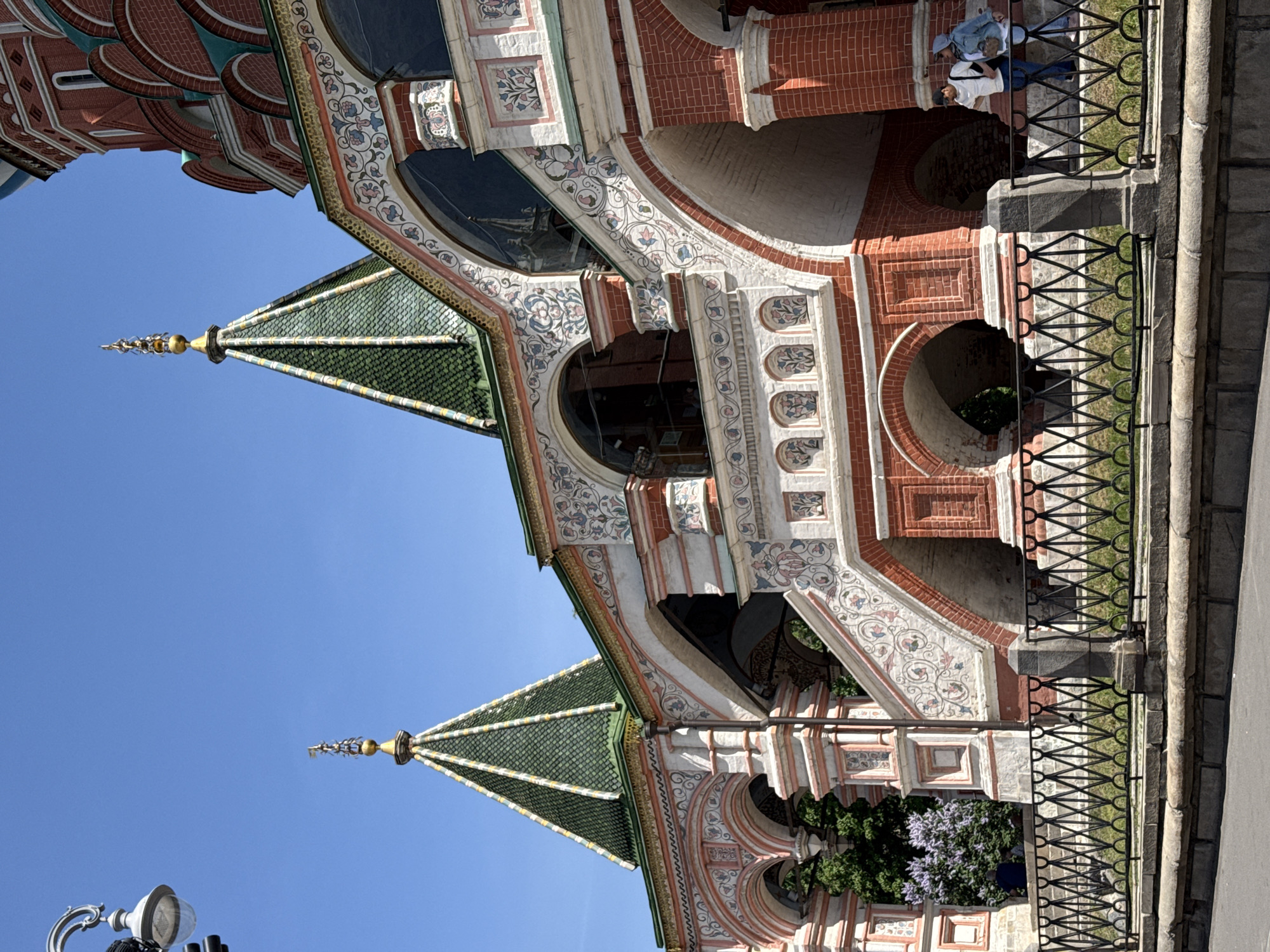 The image shows a fragment of St. Basil's Cathedral in Moscow, Russia, taken from a low angle. The view focuses on a section of the cathedral's colorful exterior, including red brick walls, ornate white decorative elements with floral motifs, and a green tiled roof with a golden spire. Two people are standing behind a black metal fence to the right of the building. The sky is clear and blue. A street lamp is visible on the left side of the image.