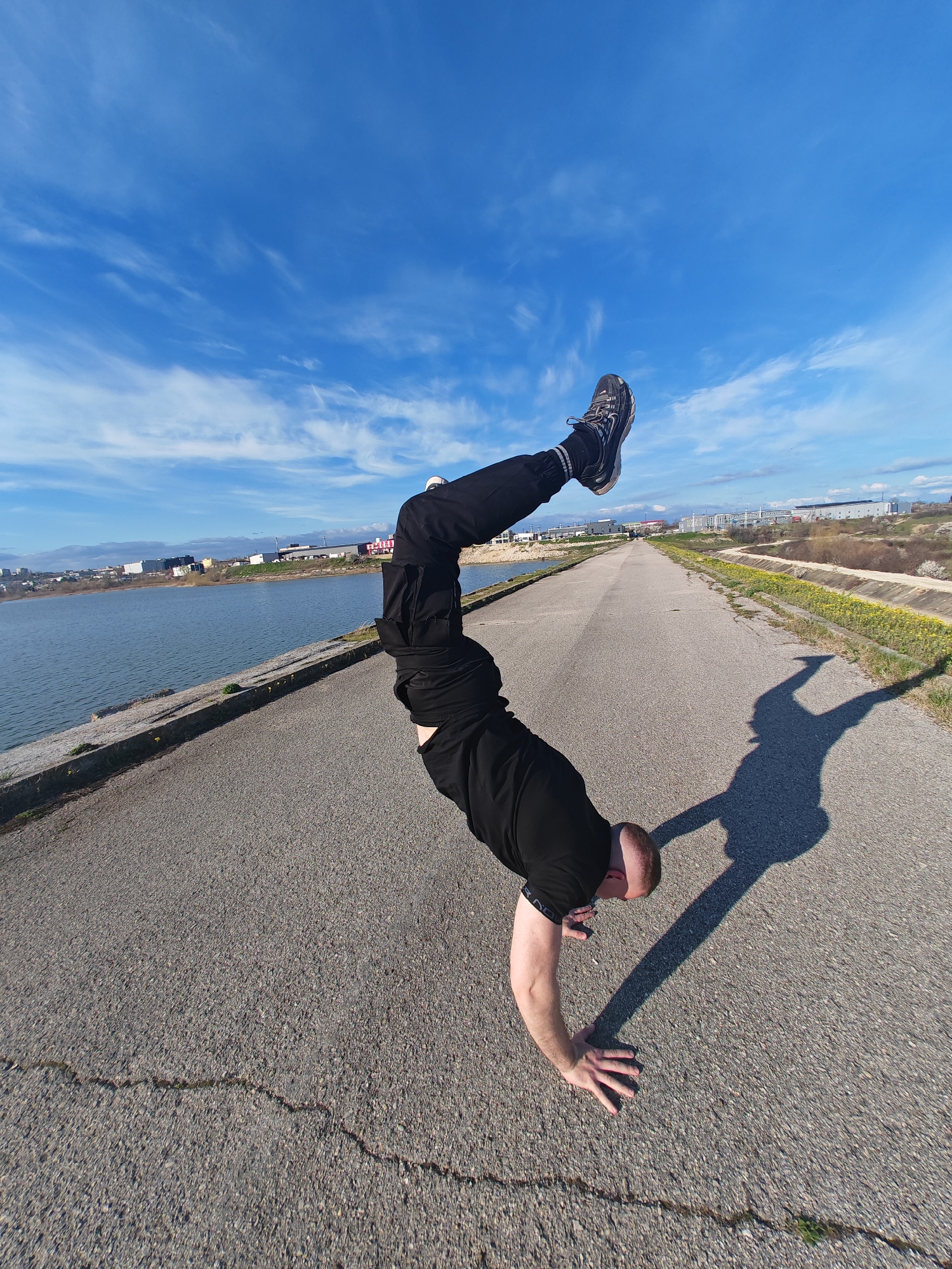 A man is performing a handstand on a paved road next to a body of water. He is wearing black pants, a black shirt, and athletic shoes. His legs are extended upward. The road is cracked in places. A shadow of the man is cast on the road. In the background, there is a body of water with buildings and greenery on the opposite shore. On the right side of the road, there is a bank with yellow flowers. The sky is blue with scattered clouds. The photograph was taken in Shumen, Bulgaria, specifically on the road next to the artificial lake "Lidl" near the Lidl store on Simeon Veliki Boulevard.