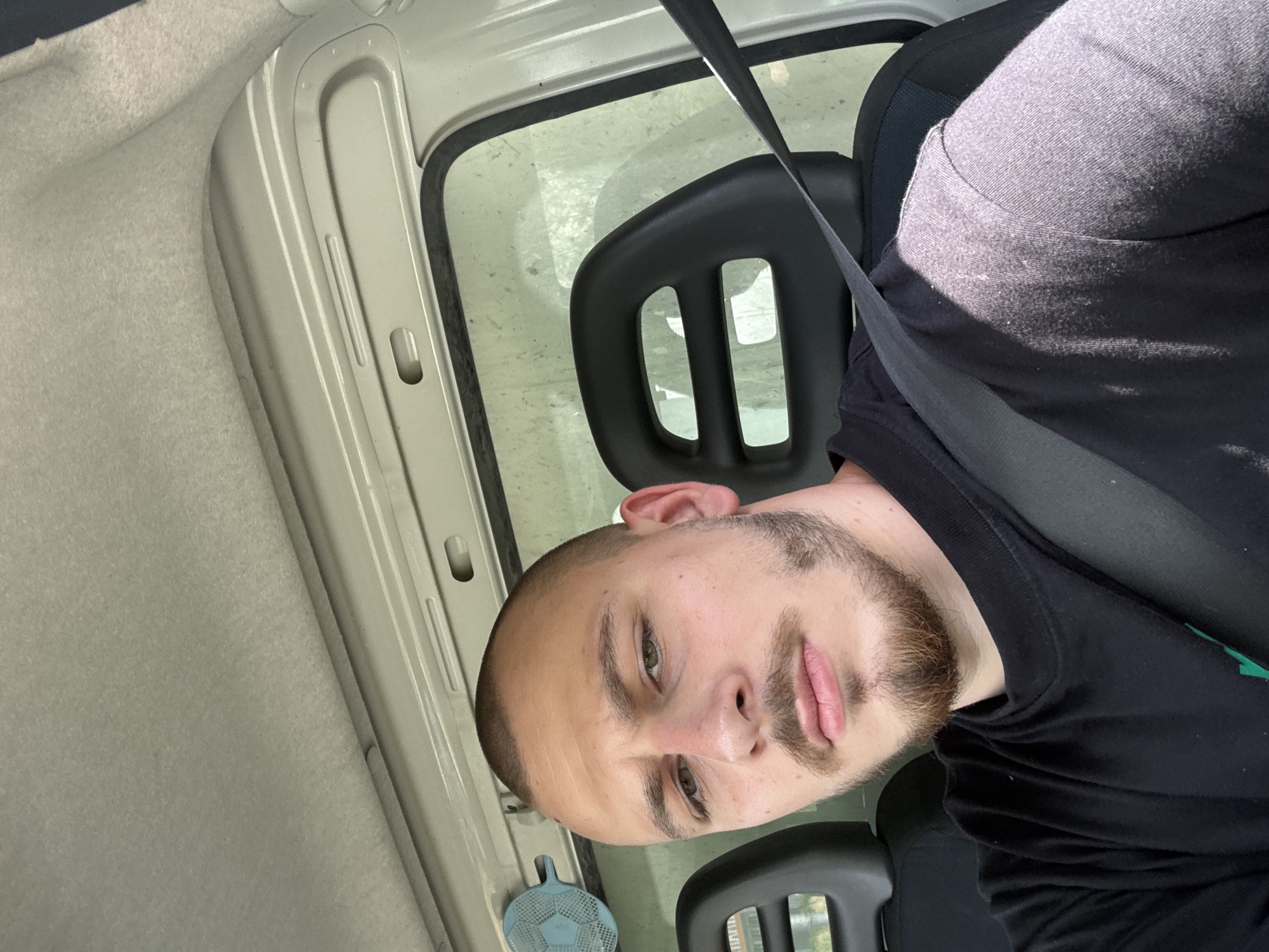 A close-up, vertical shot features a young adult male seated inside a vehicle in Kalitinovo, Bulgaria. The man, looking directly at the camera with a neutral expression, has short hair, a trimmed beard, and light-colored eyes. He wears a black t-shirt, and a black seatbelt crosses diagonally over his chest.
The setting is the interior of a car or van, indicated by the seatbelt, the design of the car seat's black headrest (which has two rectangular cut-outs), and the vehicle's light grey/beige roof lining and door frame components. A side window, visible behind the headrest, shows bright, sunlit conditions outside, though the glass appears speckled or dusty. Strong sunlight casts clear shadows, suggesting a sunny day. A small, blue, circular object with a grid pattern, possibly an air freshener or a decorative item, is attached near the bottom left of the man's head, close to the dashboard or door. There is no visible text in the image. The man appears to be taking a selfie or posing for a picture while stationary.