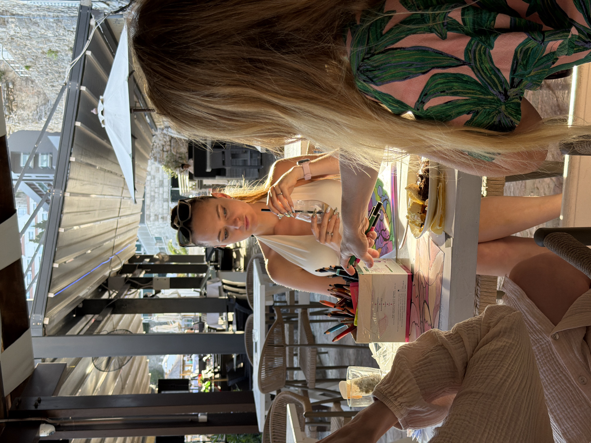A woman sits at a table at an outdoor venue in Budva, Montenegro. She is holding a glass with a straw, wearing a white top, and sunglasses on her head. Colored pencils and artwork are on the table. Two other people are partially visible at the table. The venue has an open roof structure, revealing part of the city.
