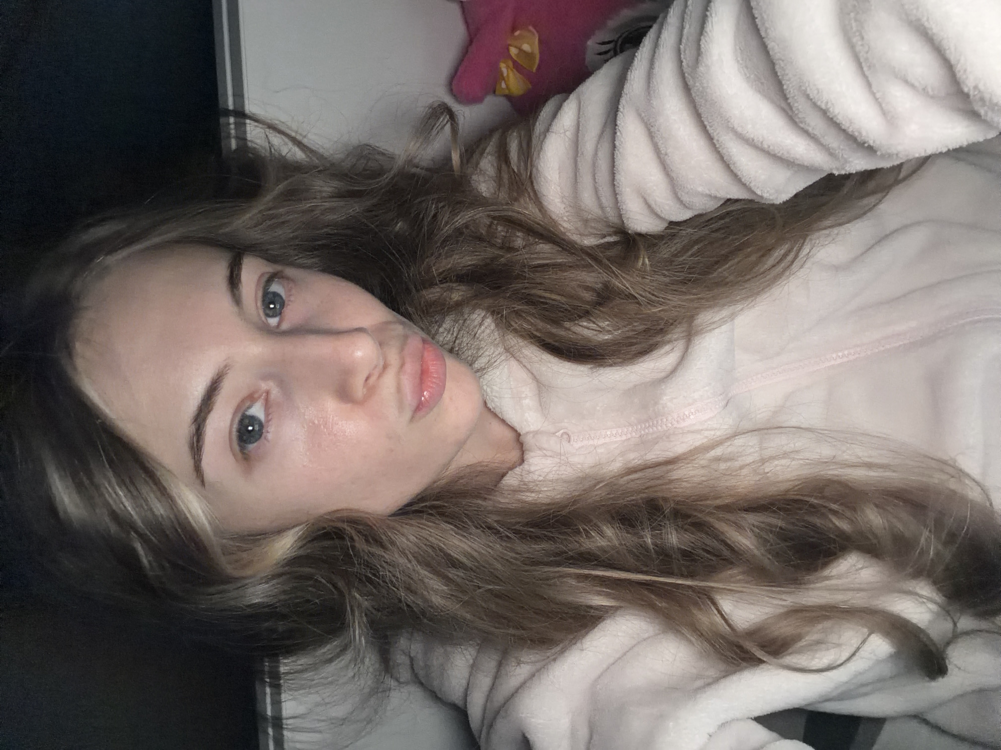A woman lies down facing the camera, with a pink stuffed animal visible near her head. She wears a light pink, fleecy top. Her hair is long, wavy, and a mix of brown and blonde. Her eyes are light blue. The background is dark and undefined. There's no evidence to suggest any location in Plovdiv, Bulgaria. The image appears to be taken indoors.
