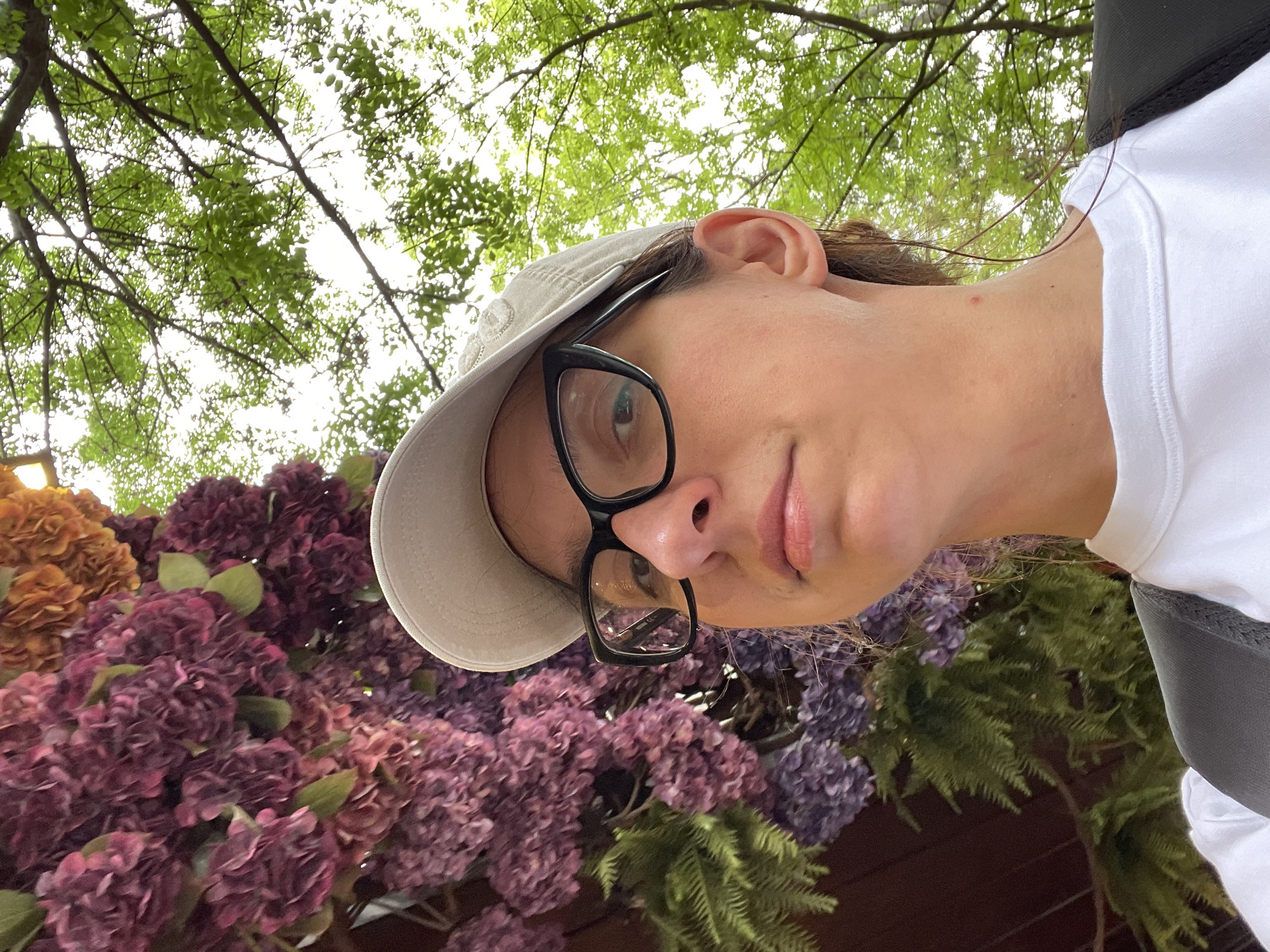 A young, fair-skinned person with glasses and a baseball cap takes a selfie from a low angle. The subject is surrounded by purple and orange flowers. Above the subject and flowers is a tree canopy with green leaves. The location is likely outdoors. It is not possible to identify the exact location in Sofia, Bulgaria, based on the visual information provided.