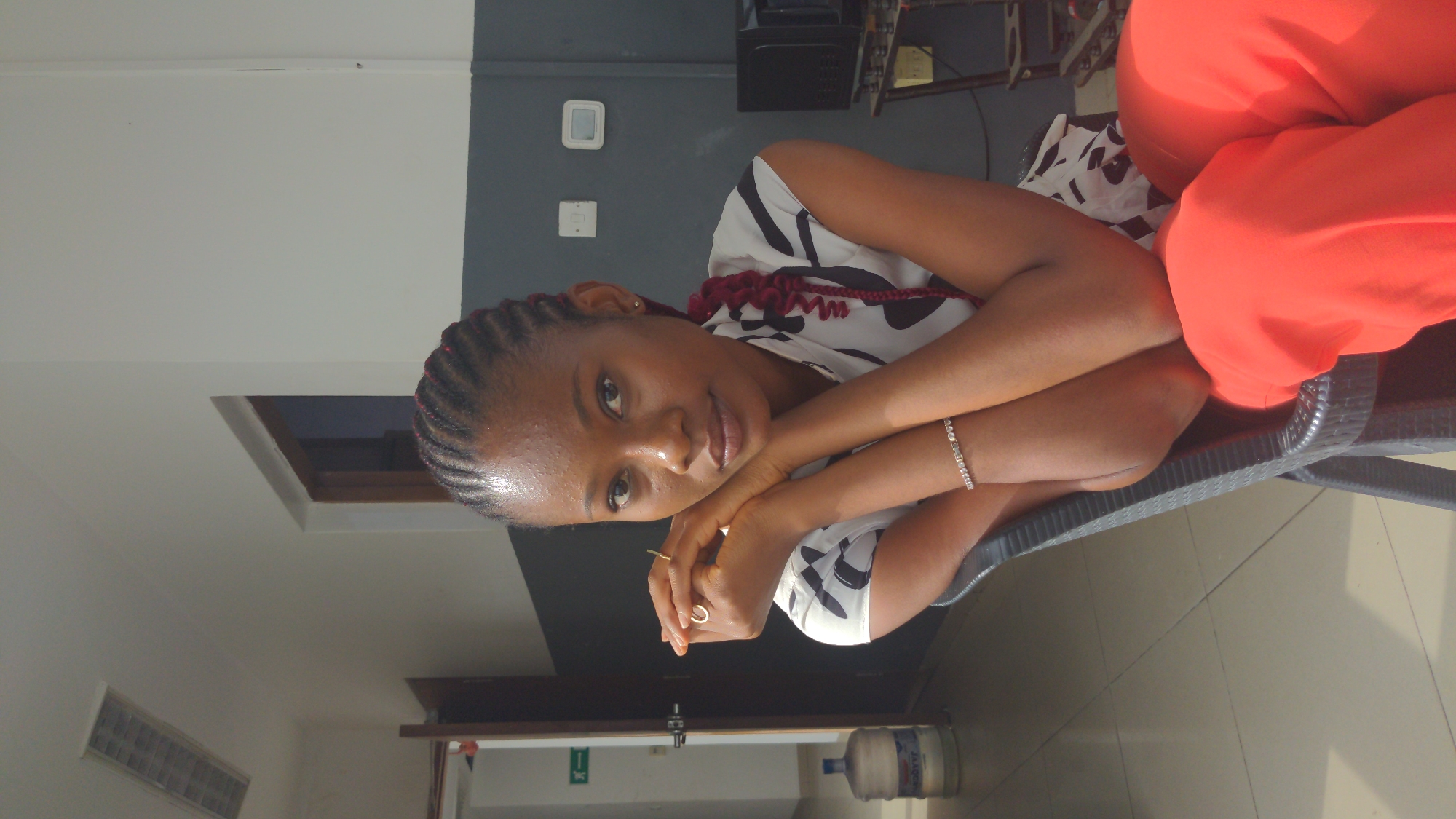 The image displays a young Black woman seated indoors in Abuja, Nigeria, looking directly at the viewer with a calm and pleasant expression. She has braided hair, featuring reddish extensions, and wears a black and white patterned sleeveless top. Her lower body is covered in bright orange fabric, possibly a skirt or trousers. She is leaning forward, resting her chin on her clasped hands. A silver-colored bracelet adorns her left wrist, and a gold-toned ring is visible on her right hand.
The setting appears to be a functional indoor space, perhaps an office, waiting room, or common area. The walls are two-toned, with a white upper section and a dark grey lower section. A rectangular window or opening is seen in the upper wall. The floor is covered with light-colored tiles. In the background, elements include a dark grey, woven-textured chair (on which the woman is seated), indistinct electronic equipment, and two white control panels or thermostats mounted on the dark grey wall. Near a doorway in the lower left, a green sign with a white arrow indicates an exit. A large water cooler bottle, with partially visible text appearing to be "Sabre" or "Sabrev," rests on the floor in the lower right. The bright lighting suggests it is daytime.