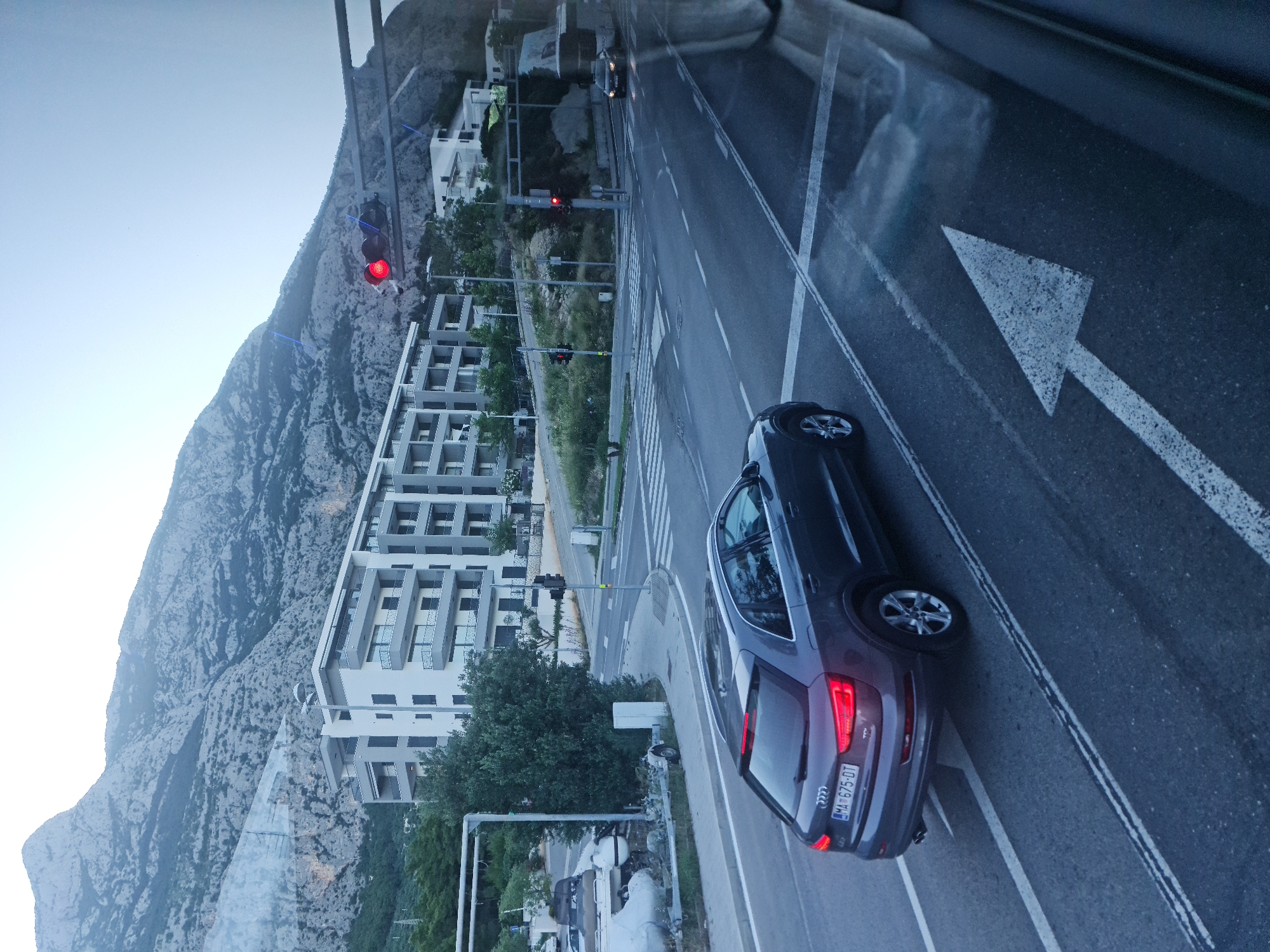 The image captures a view from a vehicle looking out at a street scene in Makarska, Croatia. In the foreground, the rear of a dark gray Audi SUV with license plate "MA 675-DT" is visible, turning into a road with a directional arrow painted on it. The SUV's taillights are illuminated. Further down the road, traffic lights are red, indicating a stop. Across the intersection, a white bus and a dark car are approaching. In the background, modern apartment buildings with balconies are situated against a backdrop of a large, rocky mountain. The sky is overcast and light blue. The scene suggests a daytime setting with moderate traffic flow.