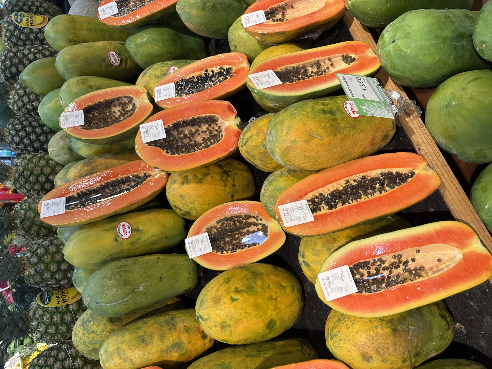 The image shows a display of halved papayas and pineapples at a grocery store. The papayas, some whole and some cut in half, have orange flesh with black seeds. Price tags are visible on many of the fruits. The pineapples are stacked on the left side of the display. The environment appears to be a produce section within a supermarket. The text found on the fruits and related signs says that the location of the image is Kuta Selatan, Indonesia.