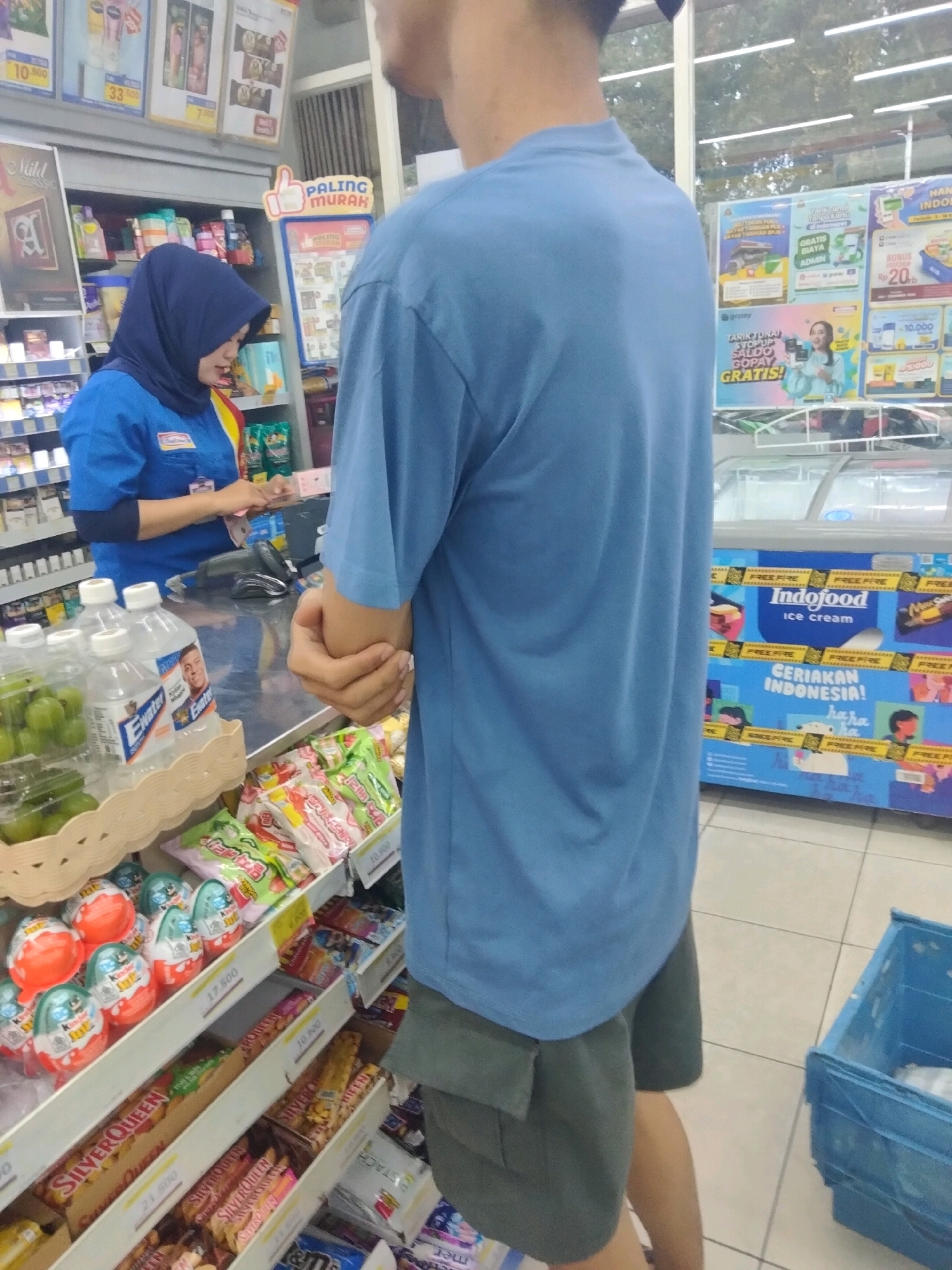 The image depicts an indoor convenience store, likely an Indomaret, in Pisangan Jaya, Indonesia. A woman wearing a blue uniform and a blue hijab is standing behind a counter, interacting with a customer who is not fully visible. The counter has a cash register and various products. Shelves in the background are stocked with diverse merchandise, including bottled drinks, snacks, and what appear to be chocolate eggs. In the foreground, shelves display more snacks, candies, and what appear to be children's toys. An open blue plastic crate is visible in the lower right corner. The store appears to be brightly lit with fluorescent lighting. The overall impression is a typical retail environment.