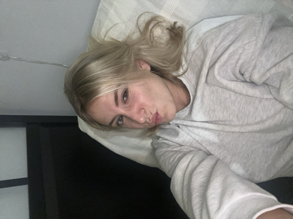 A woman with blonde hair lies on a pillow, wearing a light gray sweater. Her head is tilted, and she puckers her lips towards the camera. A black object or piece of furniture is visible on the left side of the frame, seemingly near the pillow. A thin, transparent wire runs diagonally above her head. The lighting is soft, highlighting the woman's face.