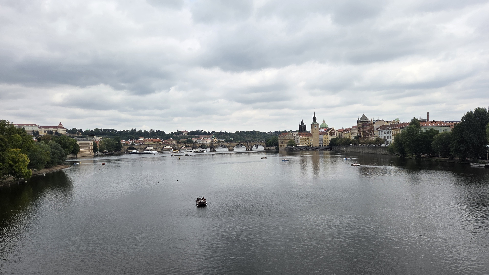 The image shows a wide, grey river flowing through the city of Prague, Czechia. On the left bank, lush green trees and a few buildings are visible. A multi-arched stone bridge, likely the Charles Bridge, spans the river in the middle ground, with several boats docked along the embankment on the far side. Further across the river, the cityscape of Prague unfolds, featuring historic architecture with red-tiled roofs and distinct clock towers, including the Old Town Bridge Tower. The sky is overcast with heavy grey clouds, and several small boats are scattered across the water. One small, dark motorboat with a couple of occupants is in the foreground. The overall scene depicts a tranquil, yet bustling, urban landscape under a cloudy sky.