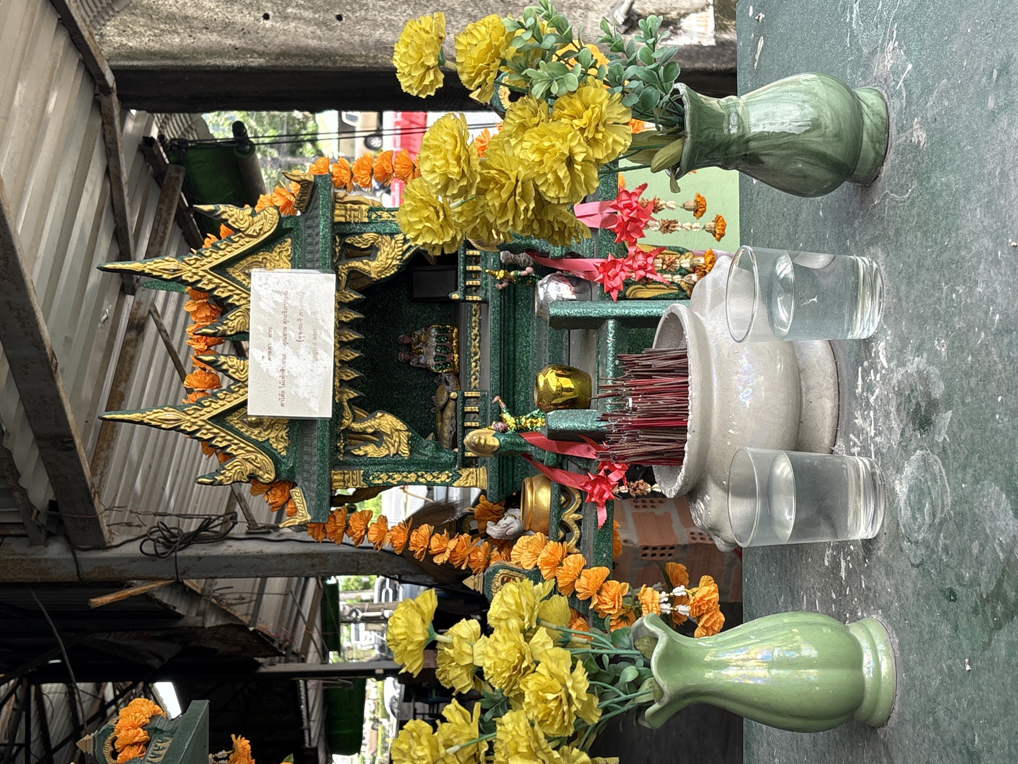 This is a medium shot of an outdoor spirit house located in Chalong, Thailand. The spirit house is ornate, with a green and gold facade decorated with orange flowers. Inside the spirit house, two small statues are visible. In front of the spirit house, there is a white ceramic bowl filled with incense sticks. Two glasses of water and two green vases filled with yellow artificial flowers flank the spirit house. The ground is a green, textured surface. In the background, there is a glimpse of a street scene with vehicles, and some structural elements of a building with a corrugated metal roof. The lighting suggests it is daytime.