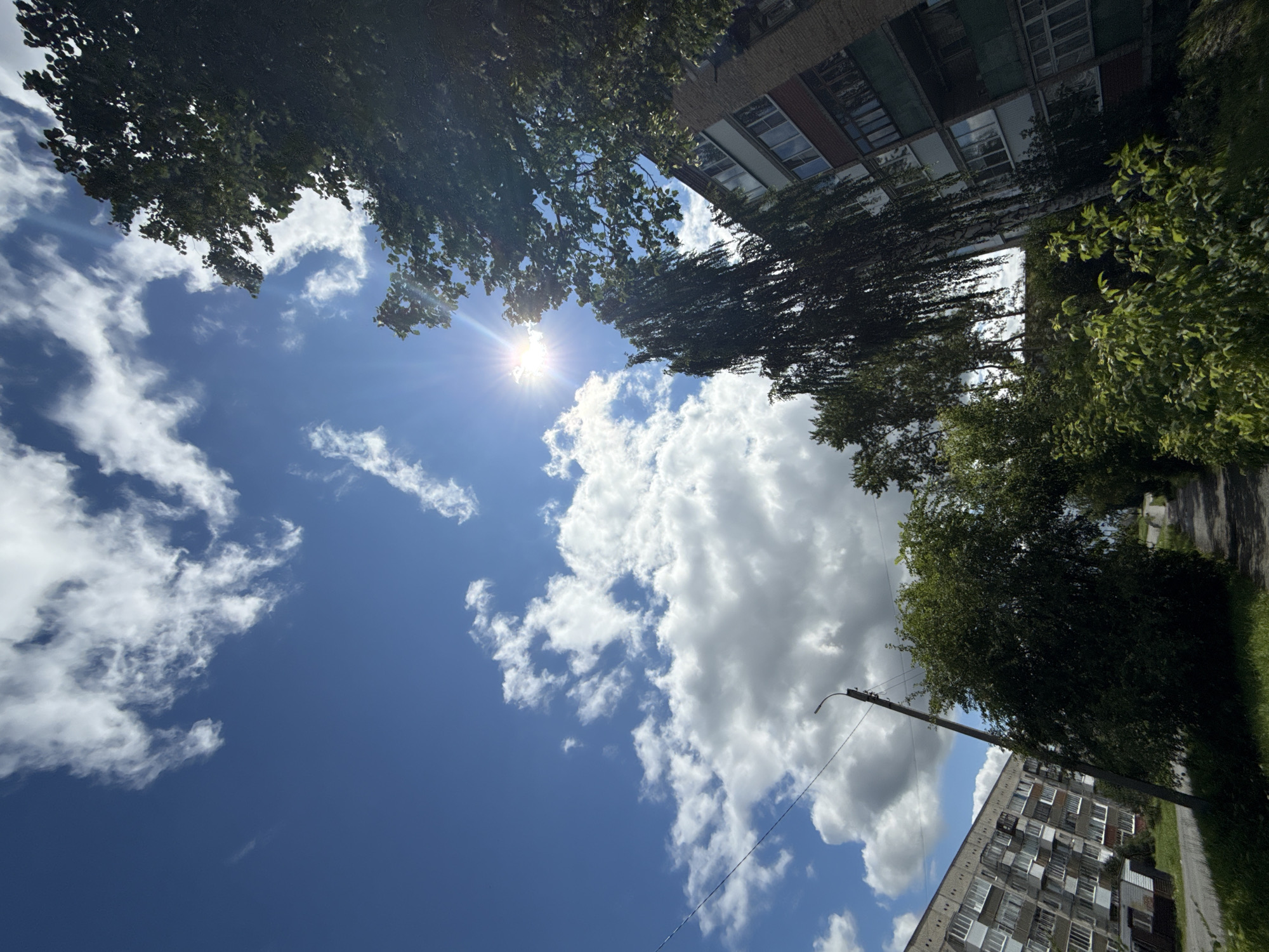 The image depicts a daytime scene in Turinsk, Russia. A bright sun shines through scattered clouds in a blue sky, creating lens flare effects. Lush green trees and vegetation are visible, with one tall tree appearing to have drooping branches. In the background, a multi-story apartment building with visible windows and balconies is present. A paved pathway or road runs through the scene, flanked by grass and trees. A utility pole with a lamp and wires is also visible. The overall impression is that of a residential area in a city on a sunny day.