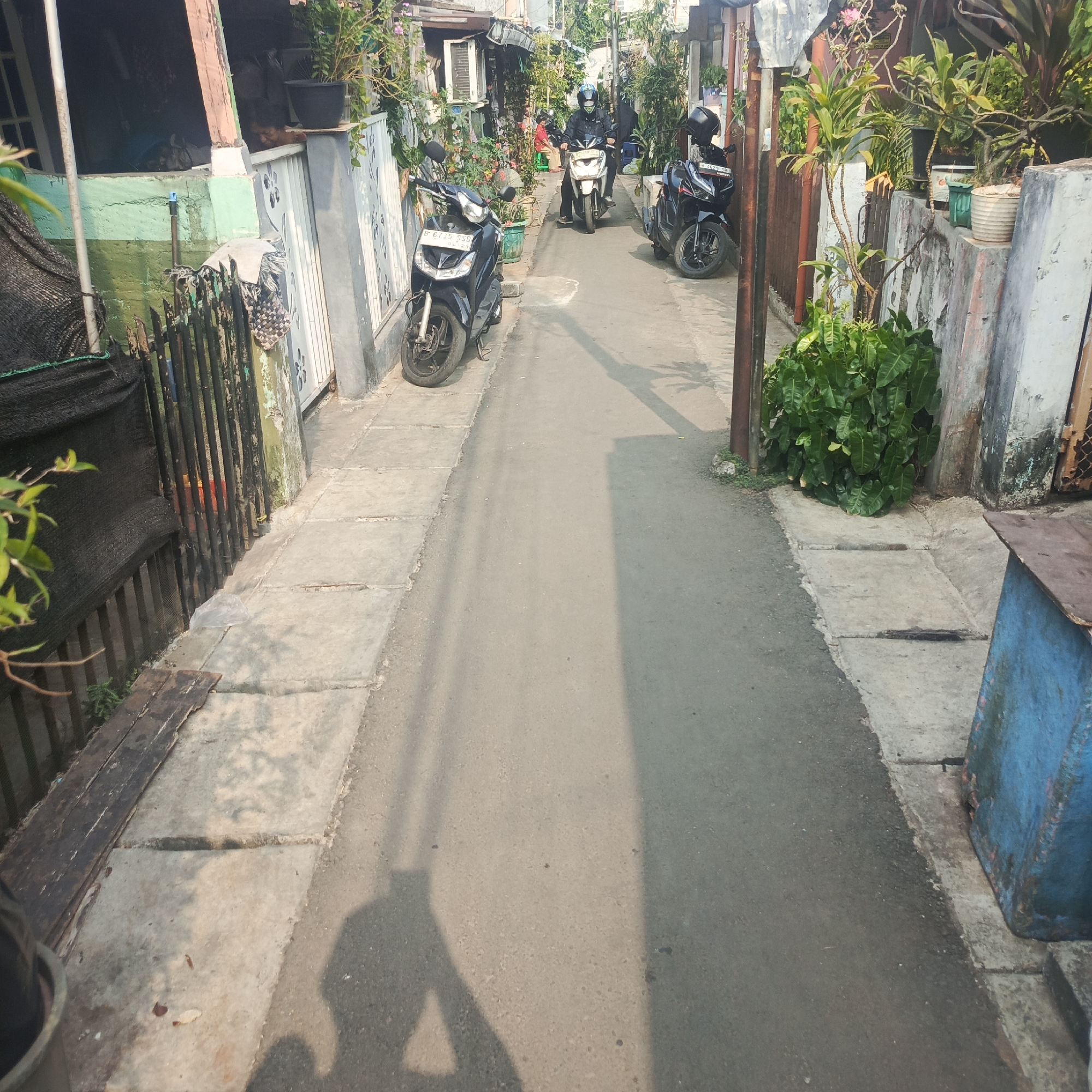 The image depicts a narrow street in a residential area, likely a kampong, in South Jakarta, Indonesia. The street is paved with asphalt and has concrete sidewalks on both sides. There are several motorcycles parked or in motion on the street. On the left side, a black scooter is parked in front of a house with a light green and white facade, a black fence, and overgrown plants. Further down the street, two more motorcycles are visible, one white and one black, with riders on them. The buildings lining the street appear to be simple houses with some greenery and potted plants. The overall environment suggests a densely populated urban residential setting.
