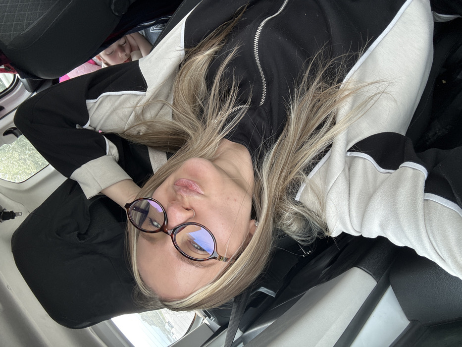 A blonde-haired female, approximately 20-30 years old, is depicted from an upward angle inside a vehicle in the city of Torbalı, Turkey. She wears dark-rimmed oval glasses and a nose stud on her left nostril. Her lips are pursed in a pouting expression, directed towards the camera. She is dressed in a black top with a silver zipper and a beige, black, and white-piped jacket. Her right arm is raised, with her hand positioned behind her head. To the right of the frame, partially obscured by the main subject and a car seat, a child's face is visible, appearing to be asleep or resting. The vehicle's interior features dark upholstery. Through the rear window, an outdoor scene is visible, showing a paved area or parking lot with distant vehicles and an indeterminate landscape under an overcast sky.