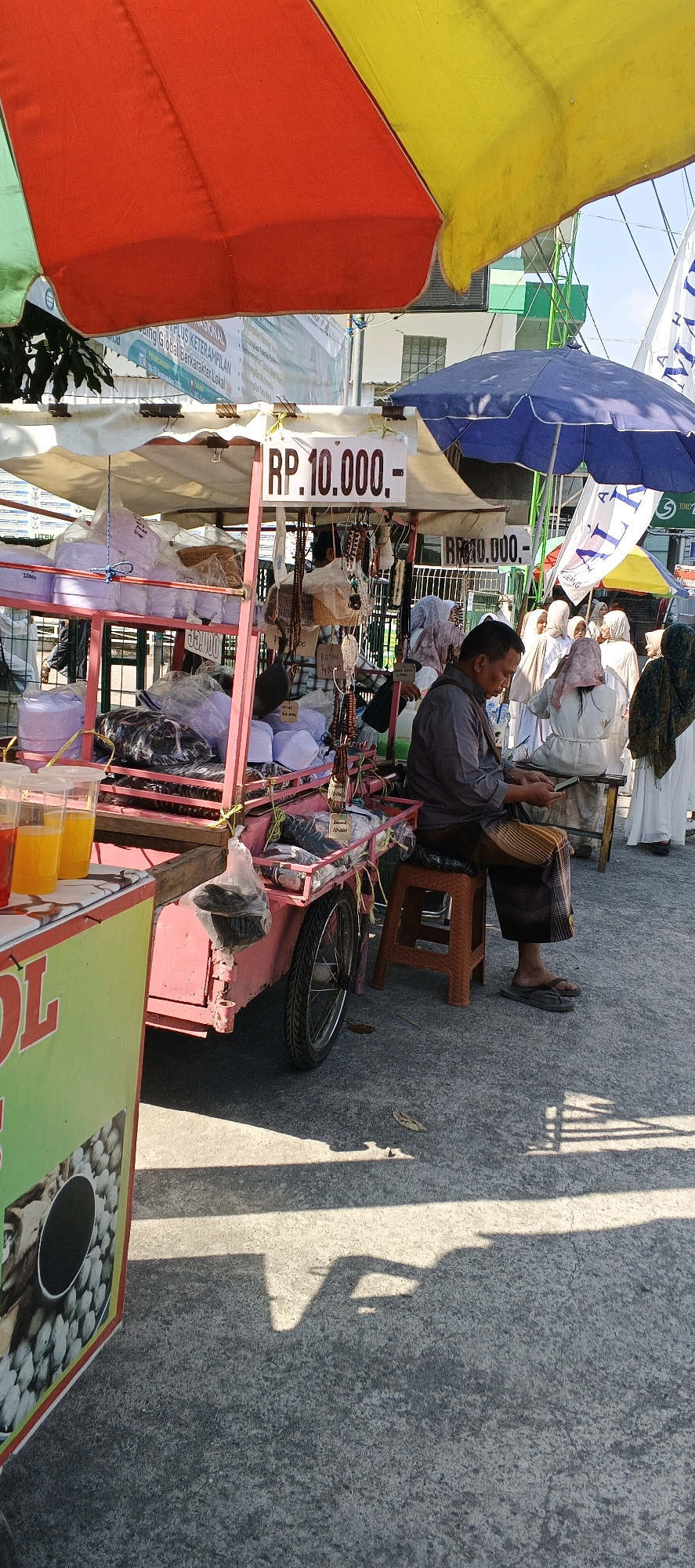 The image depicts a street scene in Sungelebak, Indonesia, with a focus on a vendor's stall. The stall is a mobile cart with a pink frame, covered by a white tarp and topped with colorful umbrellas in red, yellow, and blue. Displayed on the cart are various religious items, including white skullcaps (peci) and prayer beads, some packaged in plastic. A sign on the stall reads "RP. 10.000,-".
A man, dressed in a grey long-sleeved shirt and a patterned sarong, is seated on an orange plastic stool, looking down at a mobile phone. He is wearing flip-flops. Behind the stall, several women are gathered, all wearing white headscarves and traditional attire. They appear to be customers or passersby.
To the left of the main stall, part of another stall is visible, featuring cups of brightly colored drinks. The ground is paved with concrete, and shadows from the stalls and surrounding structures are cast across it. The overall environment suggests a bustling market or street-side shopping area.