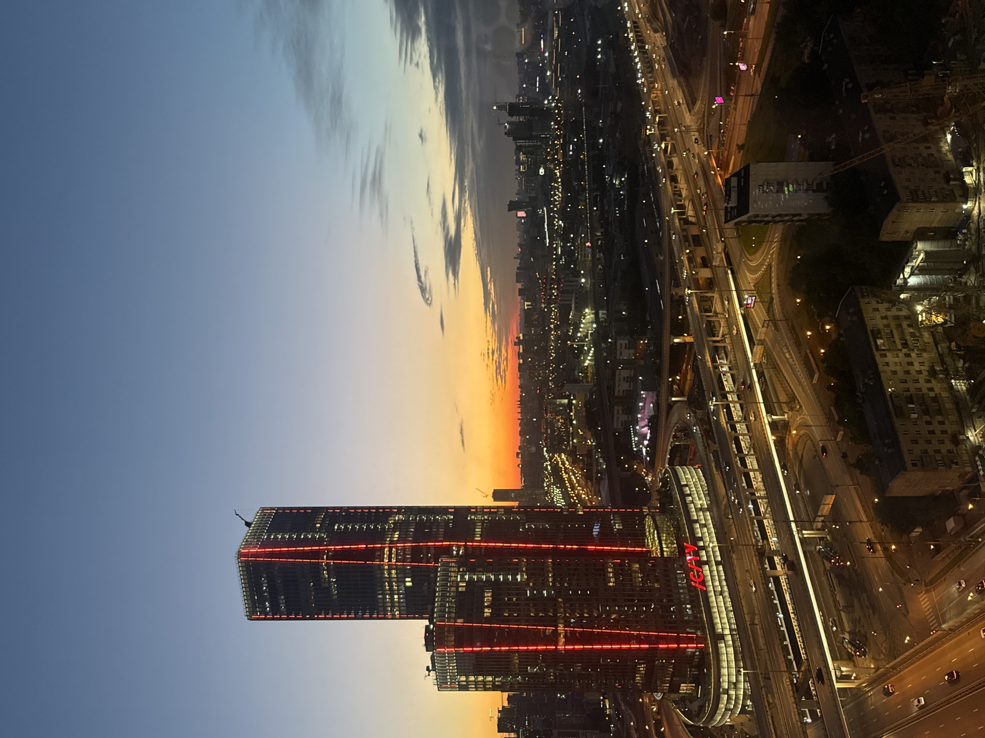 This is an aerial, twilight view of the IСITY business district in Khimki, Russia, west of Moscow. The focal point is a pair of modern skyscrapers, the taller one illuminated with vertical red lights. The lower, curved building has "ICITY" emblazoned in red lights. Below the skyscrapers, a complex network of multi-level highways and overpasses is visible, with vehicles moving along them. Several trains are also visible on elevated tracks. In the background, a sprawling cityscape stretches to the horizon, dotted with lights from other buildings and infrastructure. The sky transitions from a gradient of deep blue at the top to a warm orange and yellow glow near the horizon, indicating the sunset. Clouds are scattered across the sky, with some darker formations towards the right. The scene depicts a bustling urban environment at dusk.