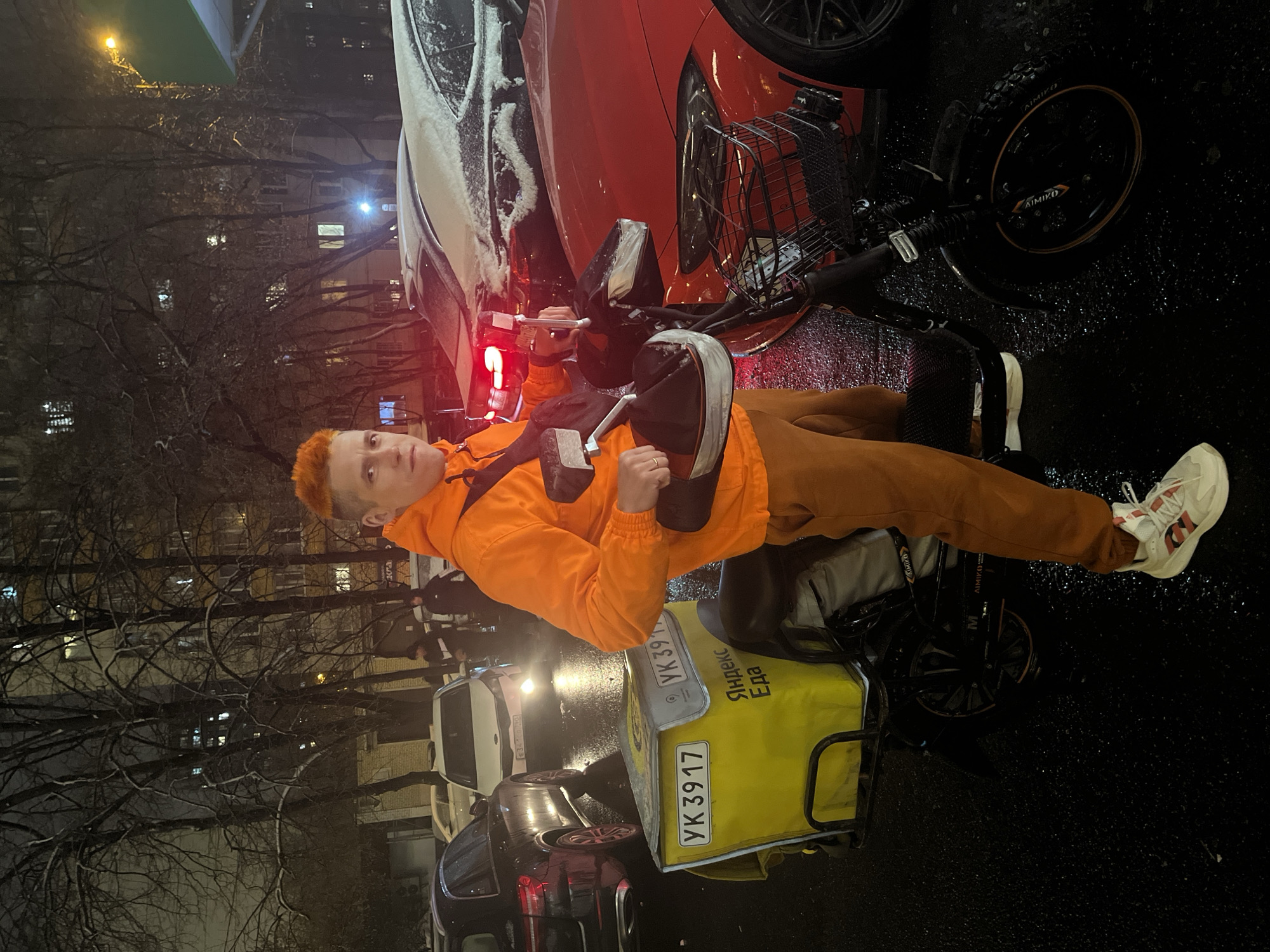 A person with orange hair is standing next to an electric bike with a yellow Яндекc Eда delivery bag on the back. The license plate on the bag reads "УК 3917". The person is wearing an orange jacket and brown pants, and white sneakers with red accents. The bike is parked near a red car covered in snow, and other cars are parked along the street. The setting is a street in Moscow, Russia, at night, with bare trees lining the road. The street is wet, suggesting recent rain or snow. Lights from buildings and streetlights illuminate the scene.