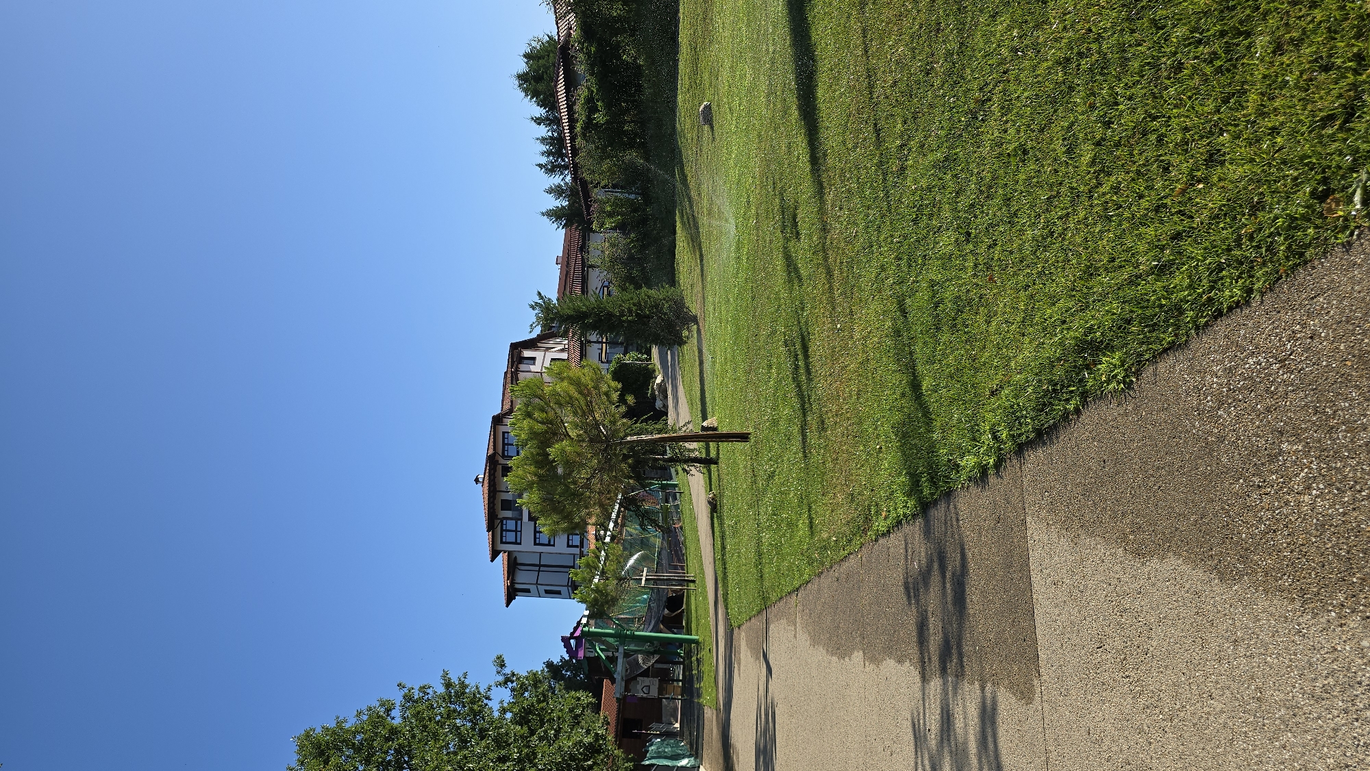 The image depicts a wide, paved walkway on the left, leading into a well-maintained grassy area. A sprinkler is actively watering the lawn in the mid-ground, creating a fine mist. A few trees are scattered across the lawn, with a larger, more mature tree in the foreground. In the background, a multi-story building with a red-tiled roof stands prominently, surrounded by lush greenery and other smaller buildings. A playground structure with nets and colorful elements is visible to the left of the main building. The sky is a clear, bright blue. The scene appears to be a resort or hotel complex in the city of Starosel, Bulgaria, on a sunny day.