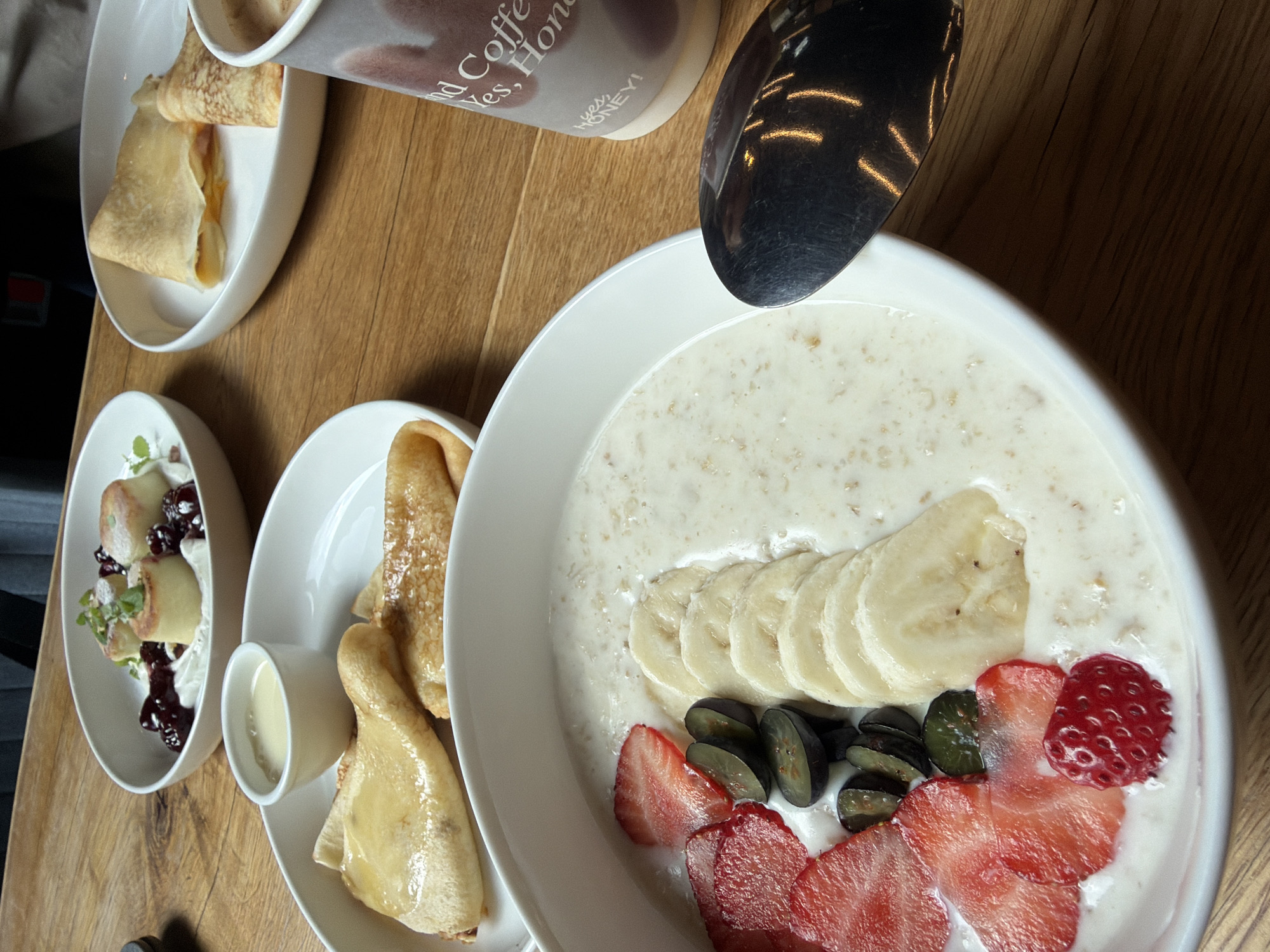 A close-up, overhead shot captures a breakfast spread on a wooden table. In the foreground, a white bowl contains oatmeal topped with sliced strawberries, blueberries, and bananas. To the right, a paper coffee cup with text that reads "and Coffee Yes, Honey!" is visible, along with a silver spoon. In the mid-ground, two white plates hold crepes, one with a creamy filling and the other with what appears to be cheese and ham. Further back, a white bowl holds a dessert with small cakes, whipped cream, and dark berries. The scene is devoid of people. The overall impression is a well-lit, organized presentation of breakfast items, likely in a cafe or restaurant setting. The city of Saint Petersburg, Russia, is not indicated by any visual cues in the image.