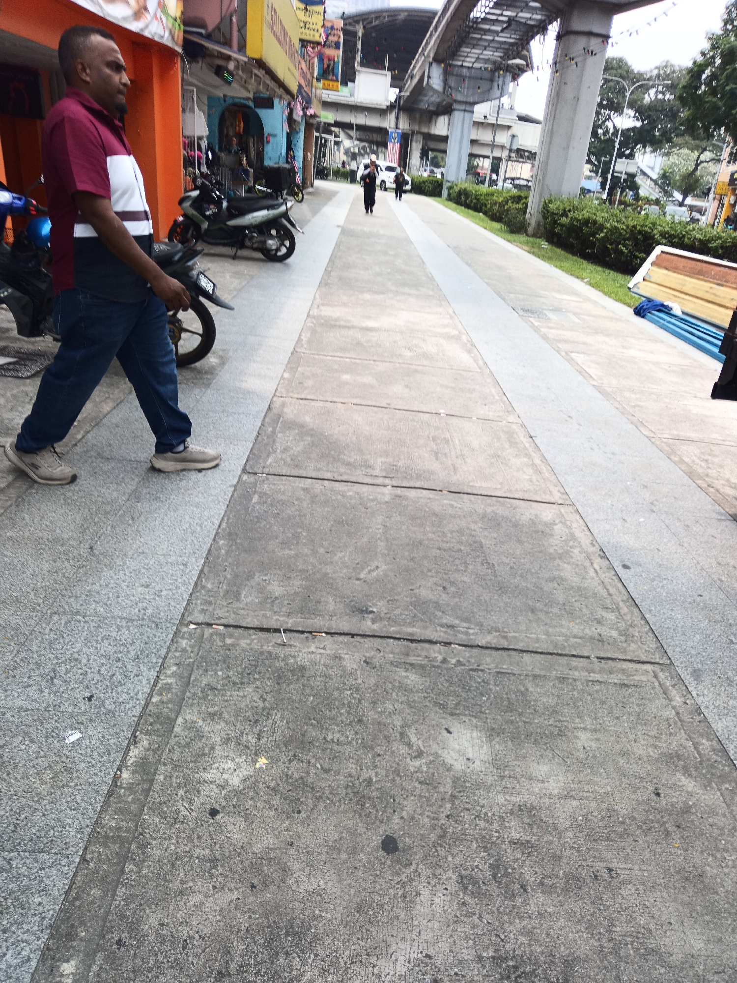 A medium shot captures a man walking on a sidewalk in Kuala Lumpur, Malaysia. The man, wearing a maroon and white collared shirt, dark blue jeans, and grey sneakers, is walking from left to right in the frame. His gaze is directed forward and slightly to the right. Behind him, parked motorcycles are visible, with one in particular being a dark-colored scooter. Further back, other pedestrians can be seen on the sidewalk, and a portion of the elevated Mass Rapid Transit (MRT) train track is visible above. The environment is urban, with buildings lining the street on the left side of the image. The sidewalk is made of concrete slabs and a contrasting granite section. The overall lighting suggests an overcast day.