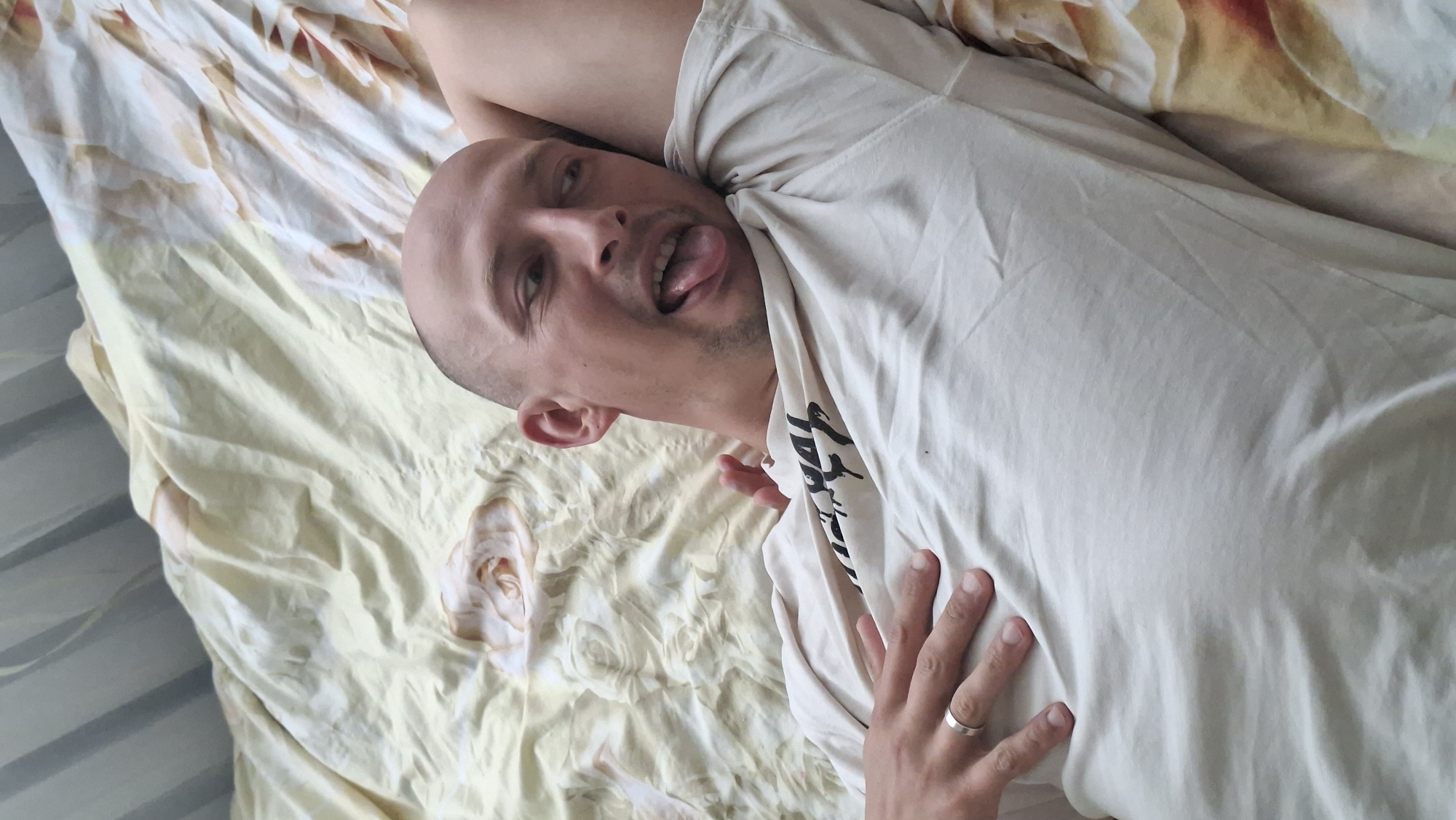 A bald man with his tongue sticking out is lying on a bed covered with a yellow and white floral patterned sheet. He is wearing a light-colored t-shirt. His right arm is bent behind his head, and his left hand is resting on his chest, wearing a ring on his ring finger. The man's eyes are looking to his right. The scene appears to be indoors, likely a bedroom. There is no indication of the city of Myadzyel, Belarus in the image.