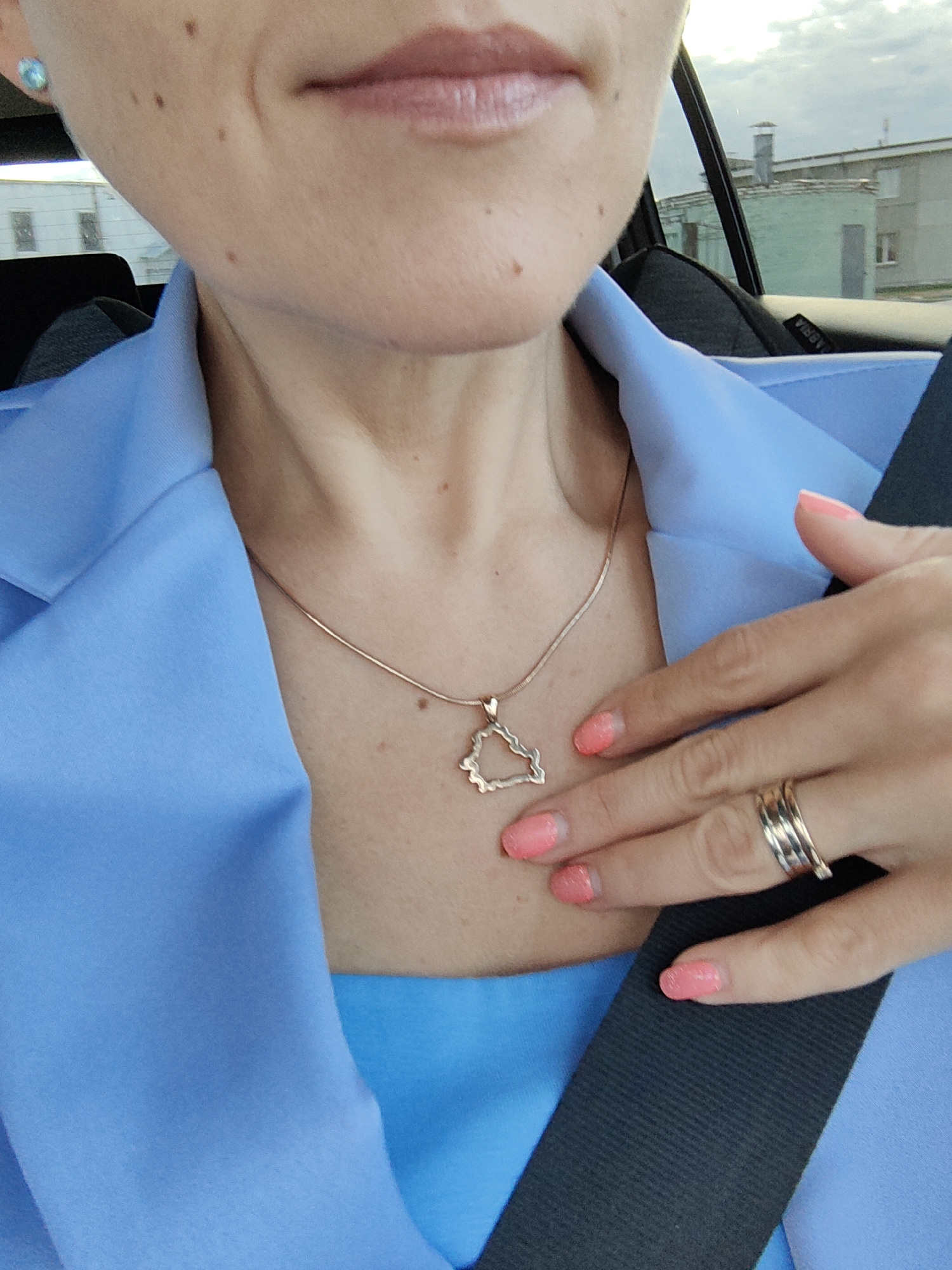 A close-up shot captures a woman's face and upper body from the chest up, inside a vehicle. She is wearing a bright blue jacket and a matching blue top. A delicate gold chain with a geometric pendant rests on her collarbone. Her right hand, adorned with coral-colored nail polish and a stacked silver ring, is resting near her neck. The background is blurred, showing a glimpse of buildings and the sky, suggesting an urban or suburban setting. The image is likely taken in Dzyarzhynsk, Belarus, based on the provided context.