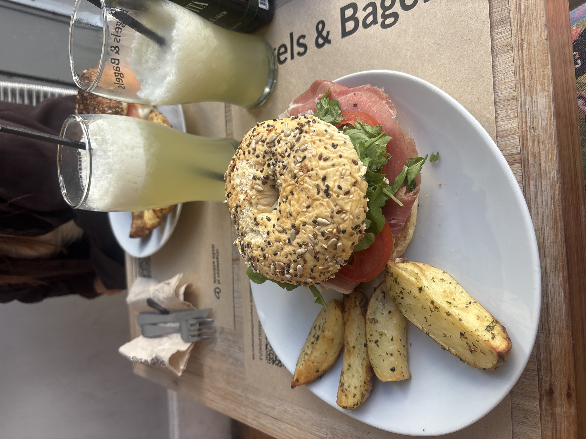 The image shows a table setting with food and drinks, likely taken in a cafe or restaurant. On the table, there's a white plate containing a bagel sandwich filled with what appears to be prosciutto, tomato, and greens, served alongside roasted potato wedges. Two tall glasses filled with a light-colored beverage with straws are also present. Cutlery and napkins are on the table next to the plate. The background hints at a wooden table surface and a printed placemat with text that includes the words "Bagels". The overall scene suggests a meal setting in a casual dining environment. There is no definitive information in the image to confirm the location in Buenos Aires, Argentina.
